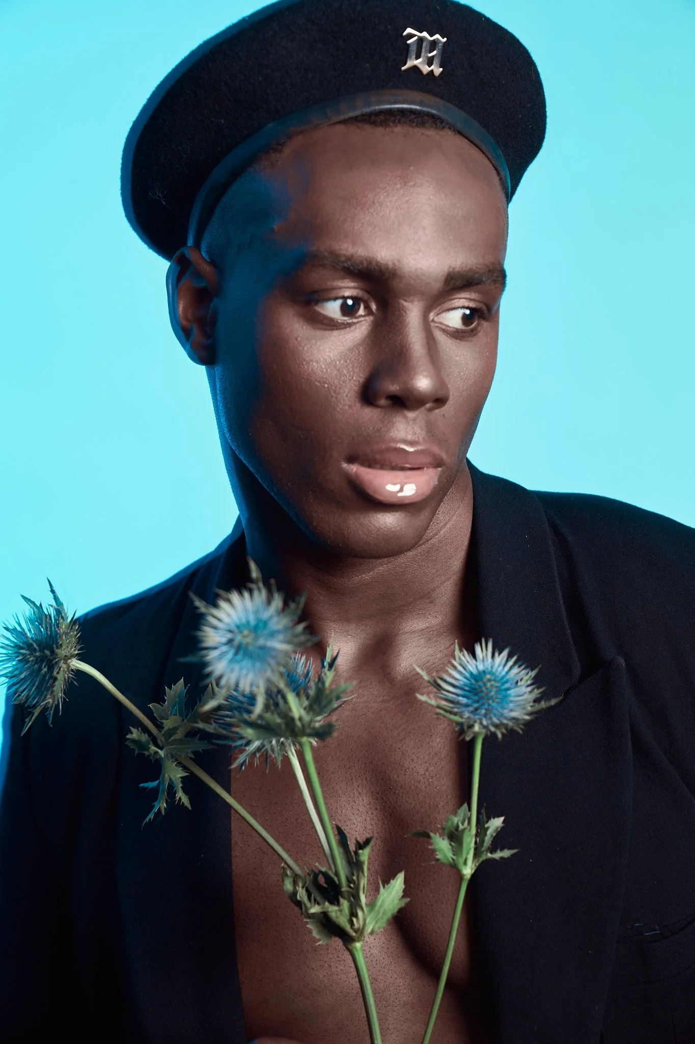 Close-up fashion portrait of a Black male model wearing a black beret and a black blazer, shirtless, holding blue thistle stalks.⁠