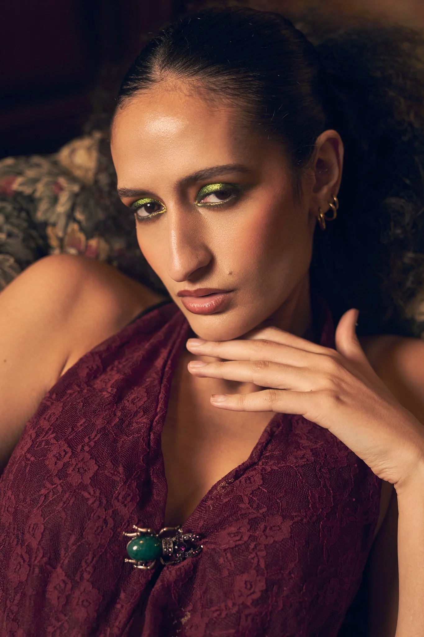 Close-up portrait of a woman wearing metallic olive eyeshadow, a textured burgundy halter top, and a green gemstone brooch.