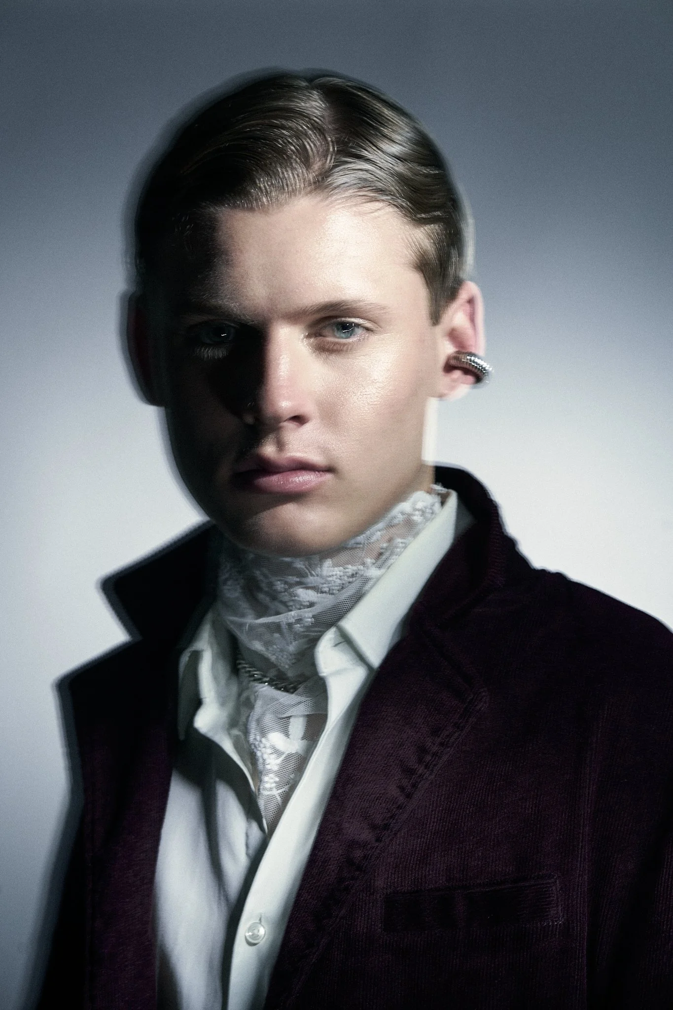 Close-up moody portrait of a man with slicked-back hair wearing a deep burgundy velvet blazer, white collared shirt, intricately detailed lace cravat, and a silver ear cuff.