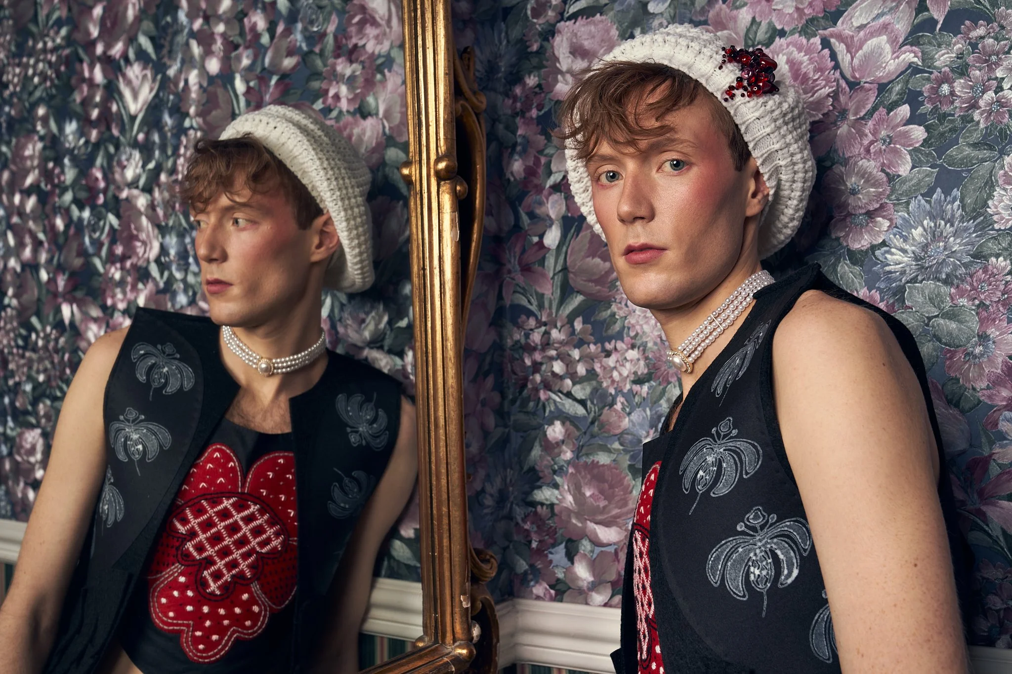 Commercial editorial portrait showing a male model wearing a knit hat, pearl choker, and dark embroidered vest, reflected in a gold vintage mirror against floral wallpaper.