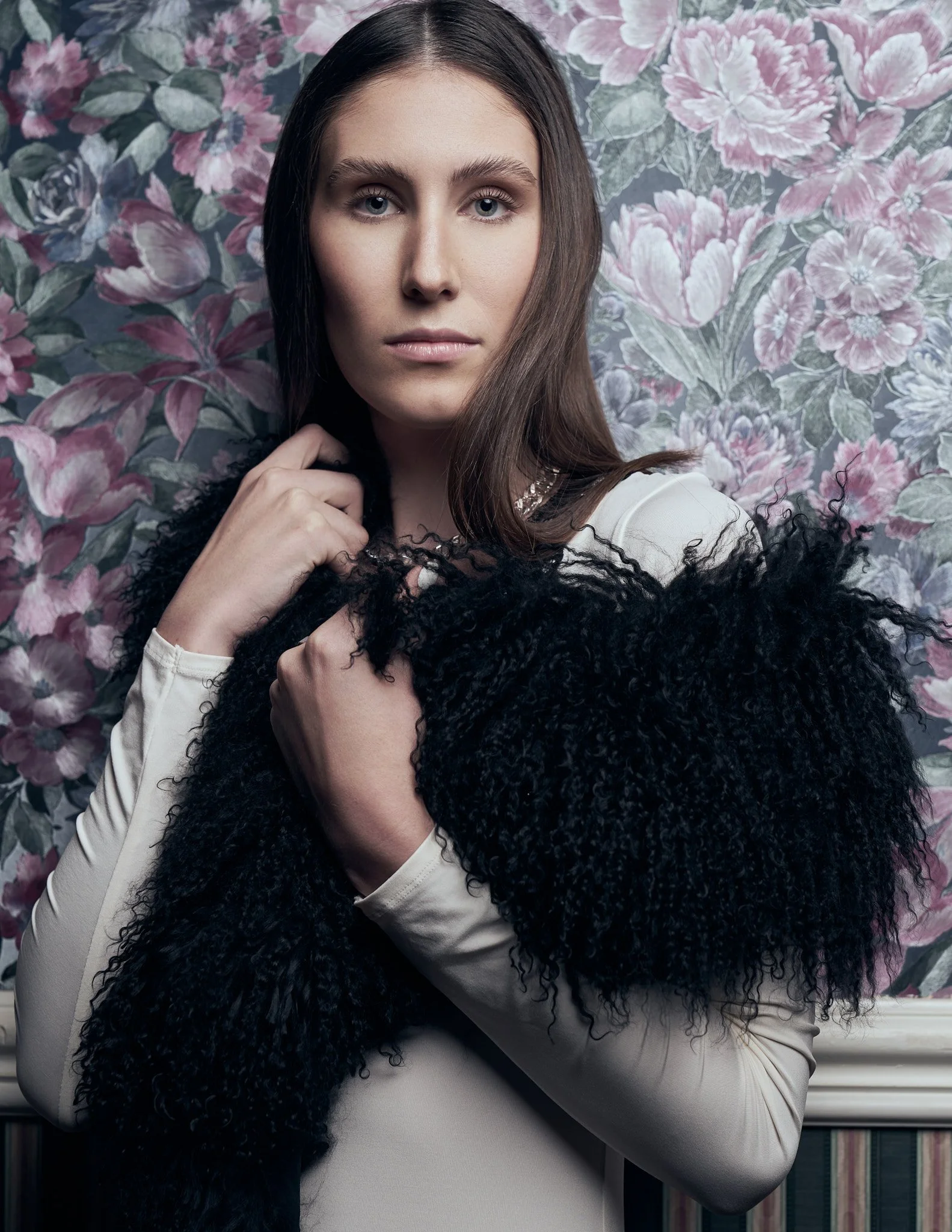 Half-length portrait of a model with dark hair wearing a long-sleeved light top wrapped in a shaggy black faux-fur stole against vintage floral wallpaper.⁠