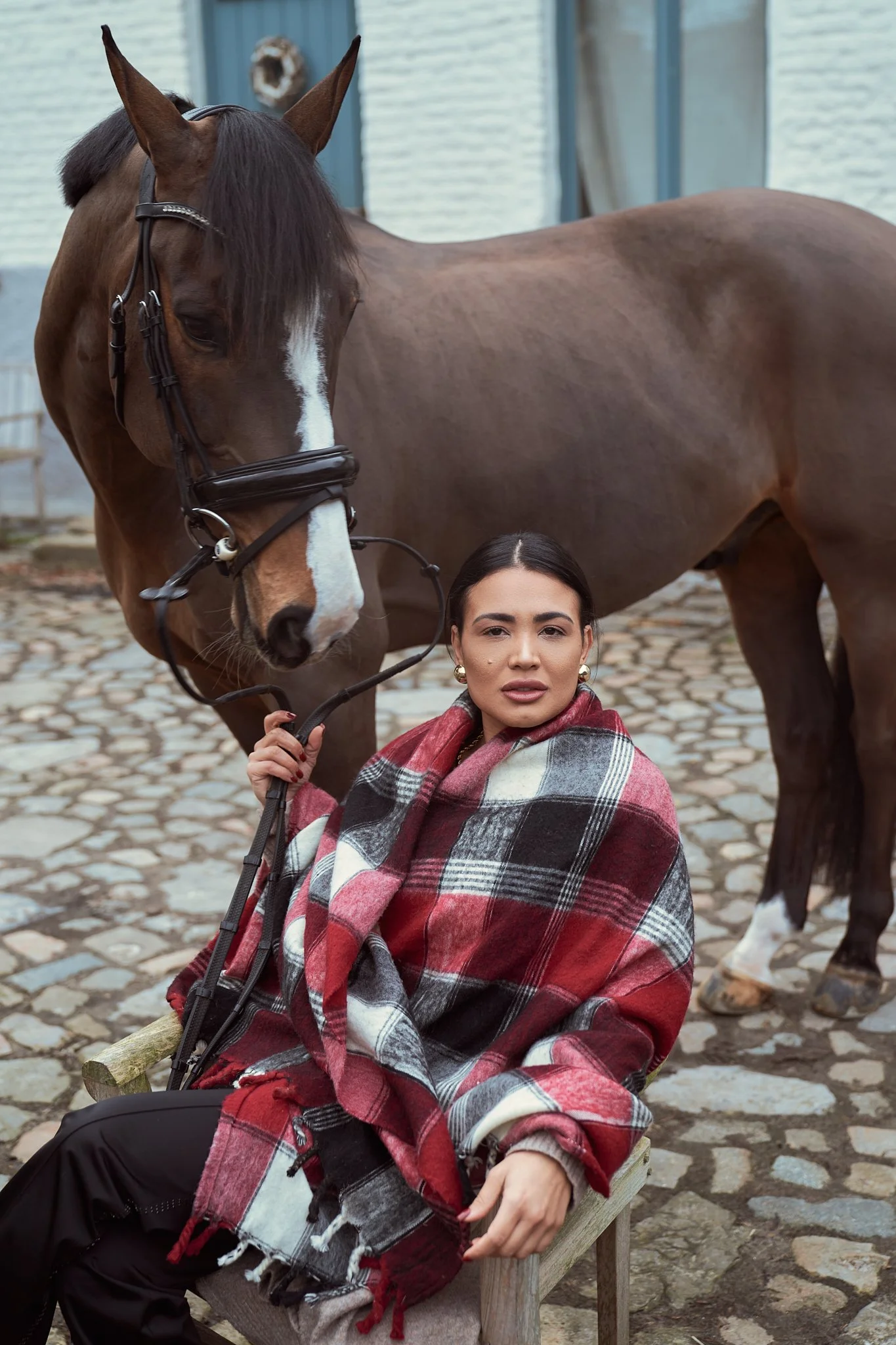 A dark-haired woman wearing a red, black, and white plaid shawl holds the reins of a large brown horse with a white face stripe outdoors on a cobblestone path.