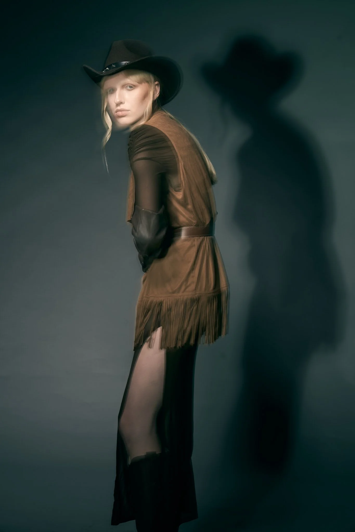 Three-quarter Western fashion shot of a blonde model wearing a cowboy hat, sheer dark sleeves, and a brown suede vest.⁠