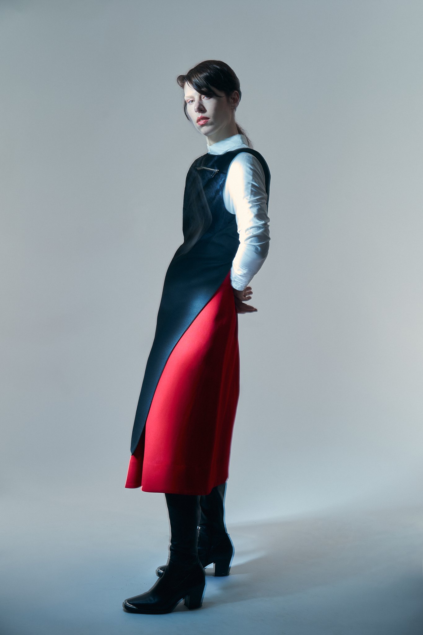 ⁠A structural fashion editorial portrait showing a side profile of a model, highlighting the asymmetrical drape and layered construction of a black tunic and a vibrant red skirt against a minimal grey background.⁠