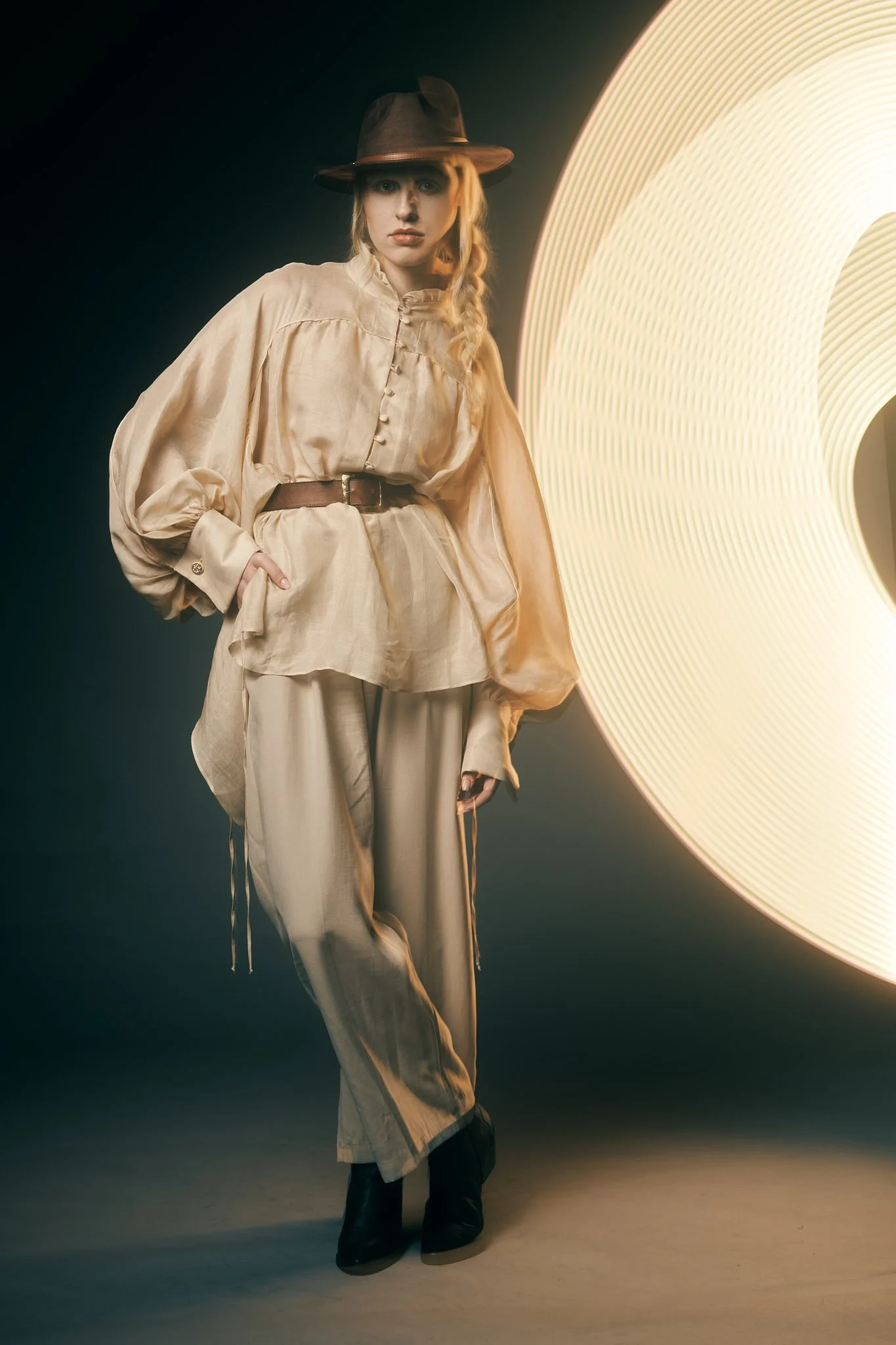 Full-length fashion portrait of a model in a wide-brim hat and high-waisted beige trouser suit standing near a large studio light modifier.⁠