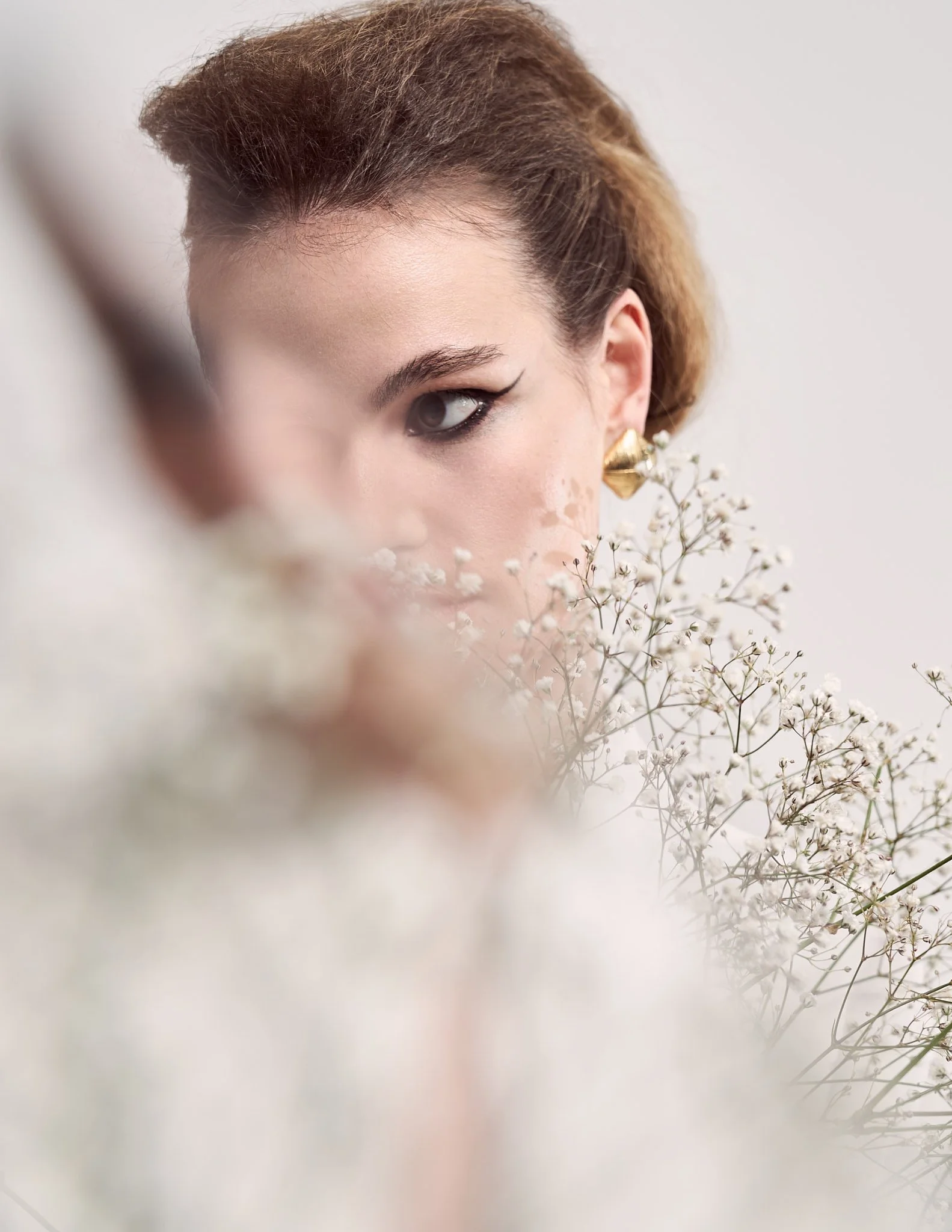 Close-up fashion beauty shot of a model with winged eyeliner and gold drop earrings framed behind soft, out-of-focus white baby's breath flowers.⁠