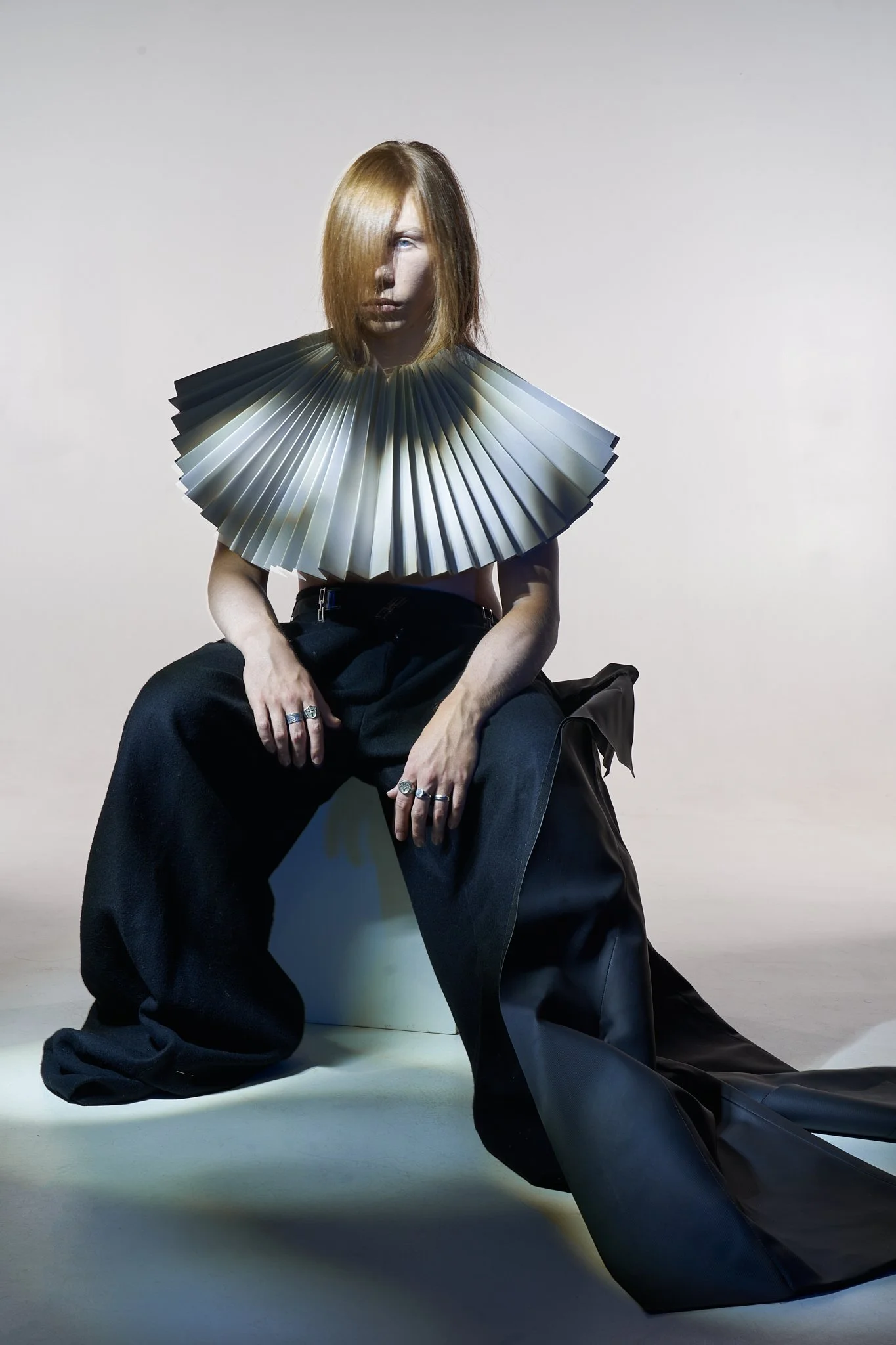 Full-body portrait of a model with long reddish hair covering half their face, wearing an accordion-pleated fan collar piece, wide-leg black trousers, and silver rings, seated against a neutral studio backdrop.