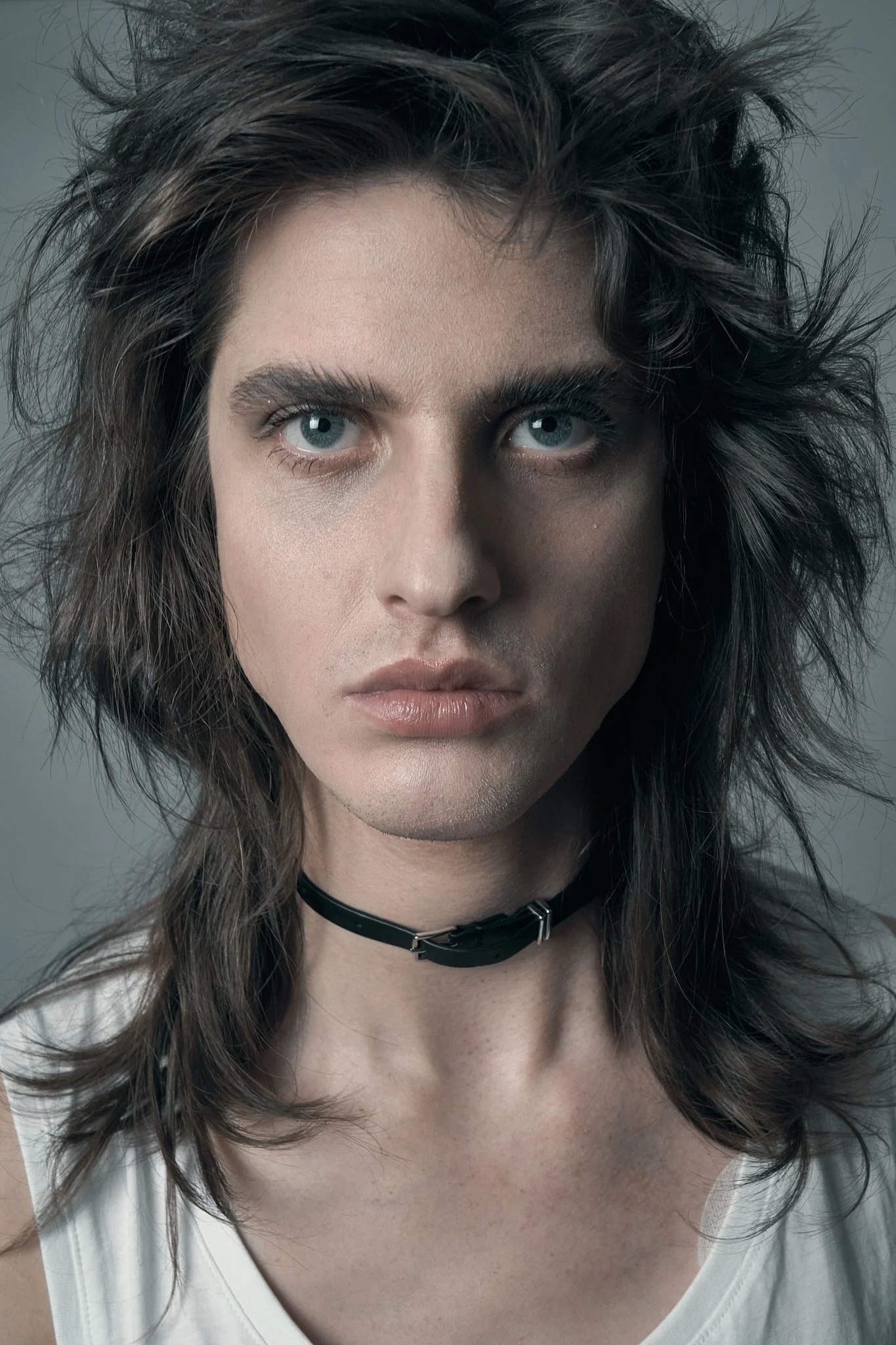 Close-up beauty portrait of a male model with layered shag hair, dramatic eye makeup, and a black choker collar.⁠