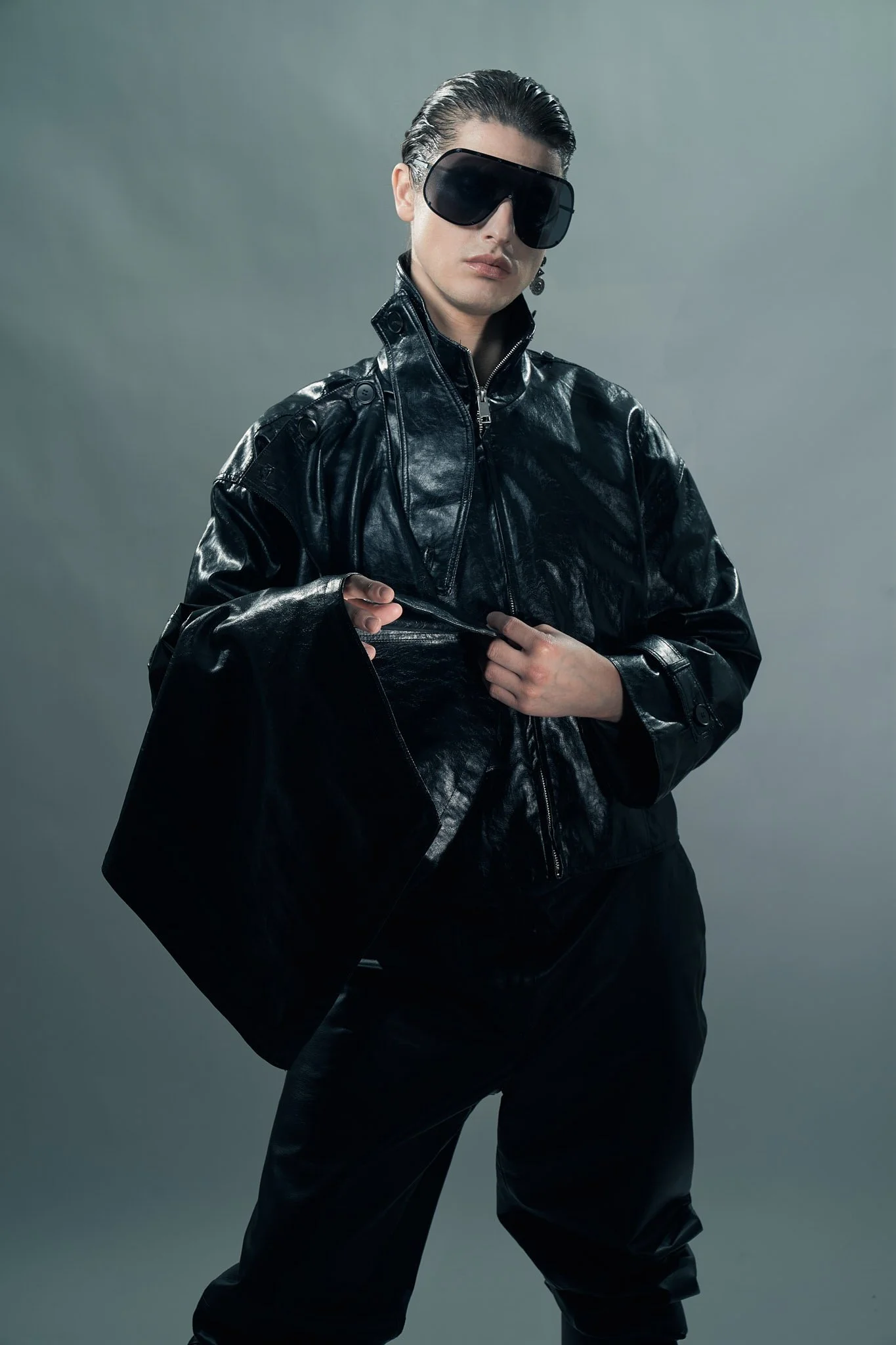 Full-length edgy menswear photograph of a model wearing oversize black shield sunglasses and a dark high-collar leather jacket.⁠