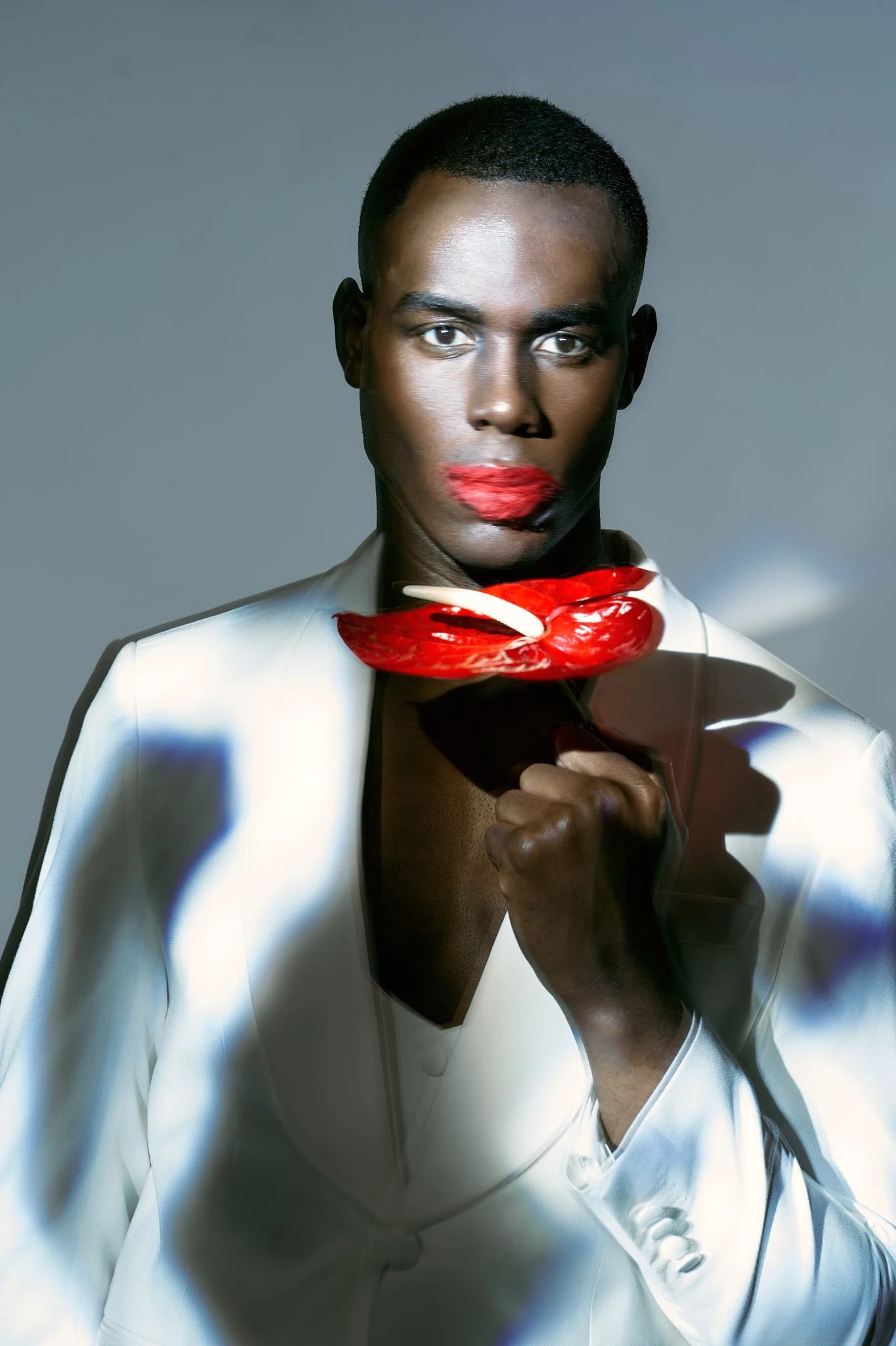 Fashion close-up of a Black male model with red lipstick, shirtless in a white blazer, holding a single red and white flower under dramatic light.⁠