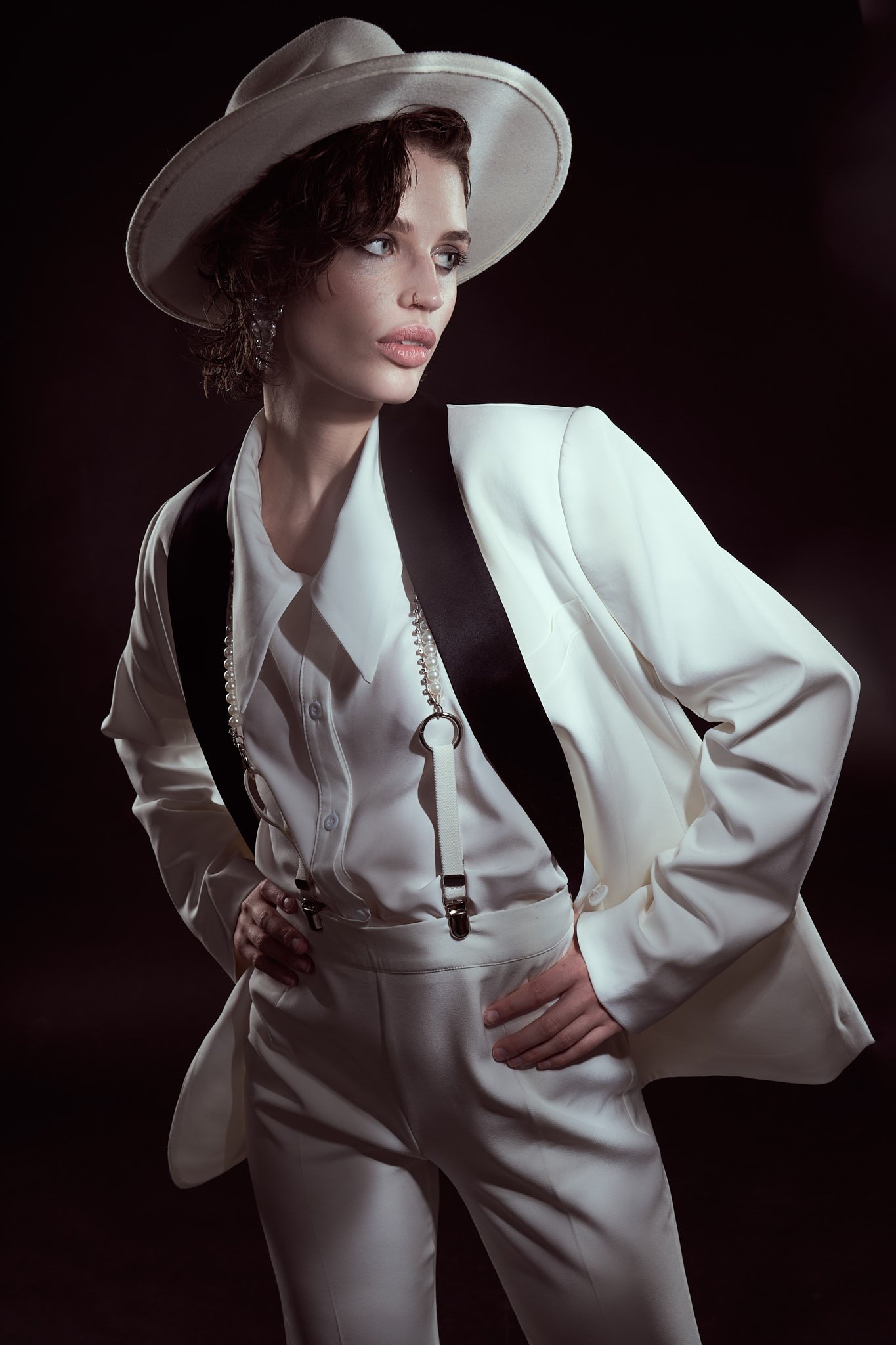 Fashion portrait of a model wearing a white suit, white fedora, and black suspenders posing against a dark backdrop.⁠