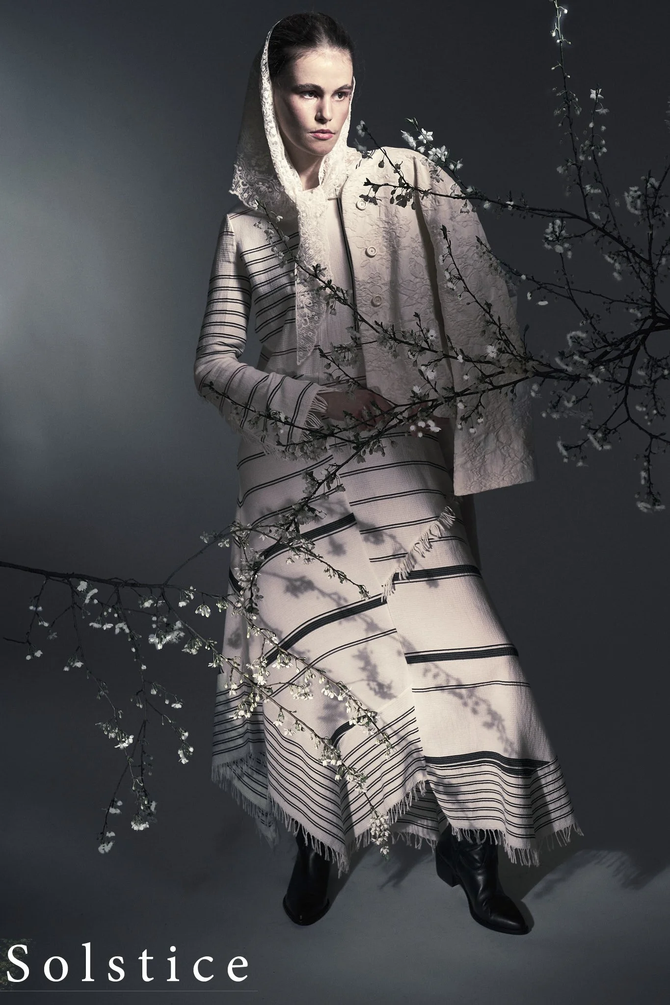Editorial fashion photography of model in striped dress and lace headscarf surrounded by cherry blossom branches, dark studio background, Simon Rogghe Ghent fashion photographer, published in Solstice Magazine