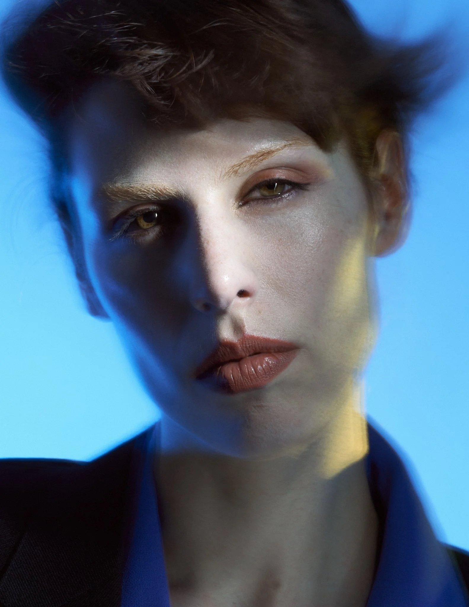 Close-up beauty shot in a blue light setting focusing on a model's face, showing natural makeup and defined features.⁠
