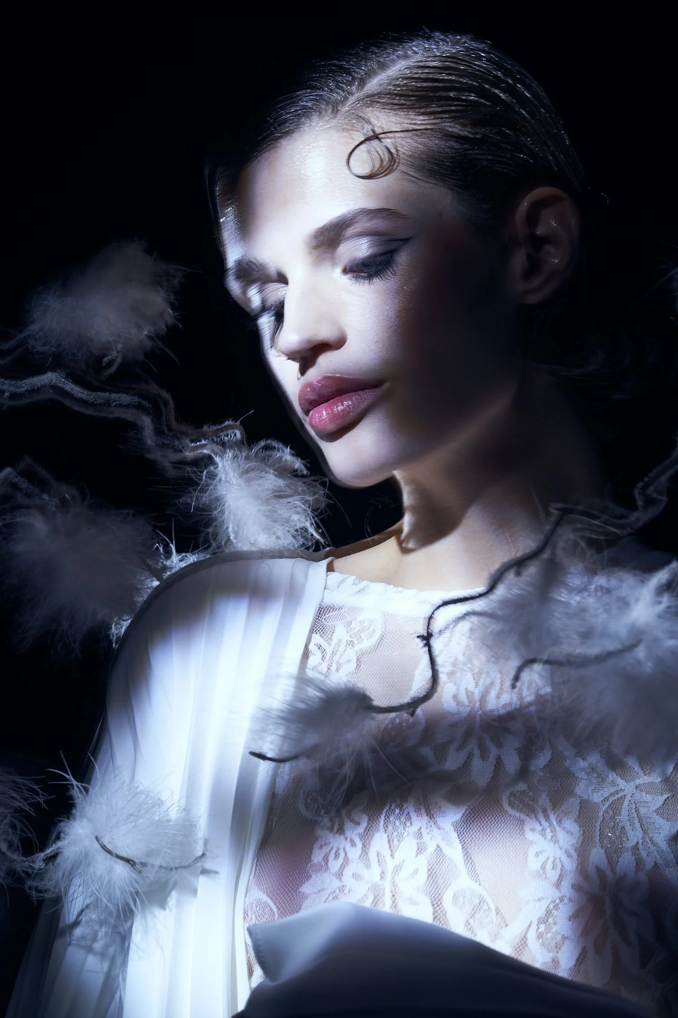 Atmospheric fashion portrait of a model in a lace top under dramatic lighting, surrounded by floating white feathers.⁠