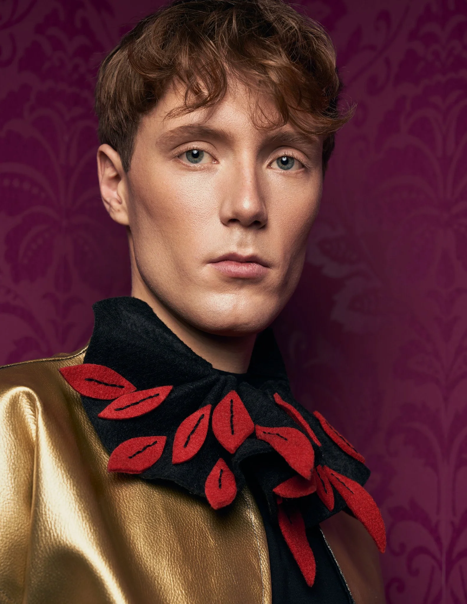 Close-up portrait of a male model wearing a metallic gold jacket with an oversized black collar adorned with cut-out red leaf appliqués.⁠