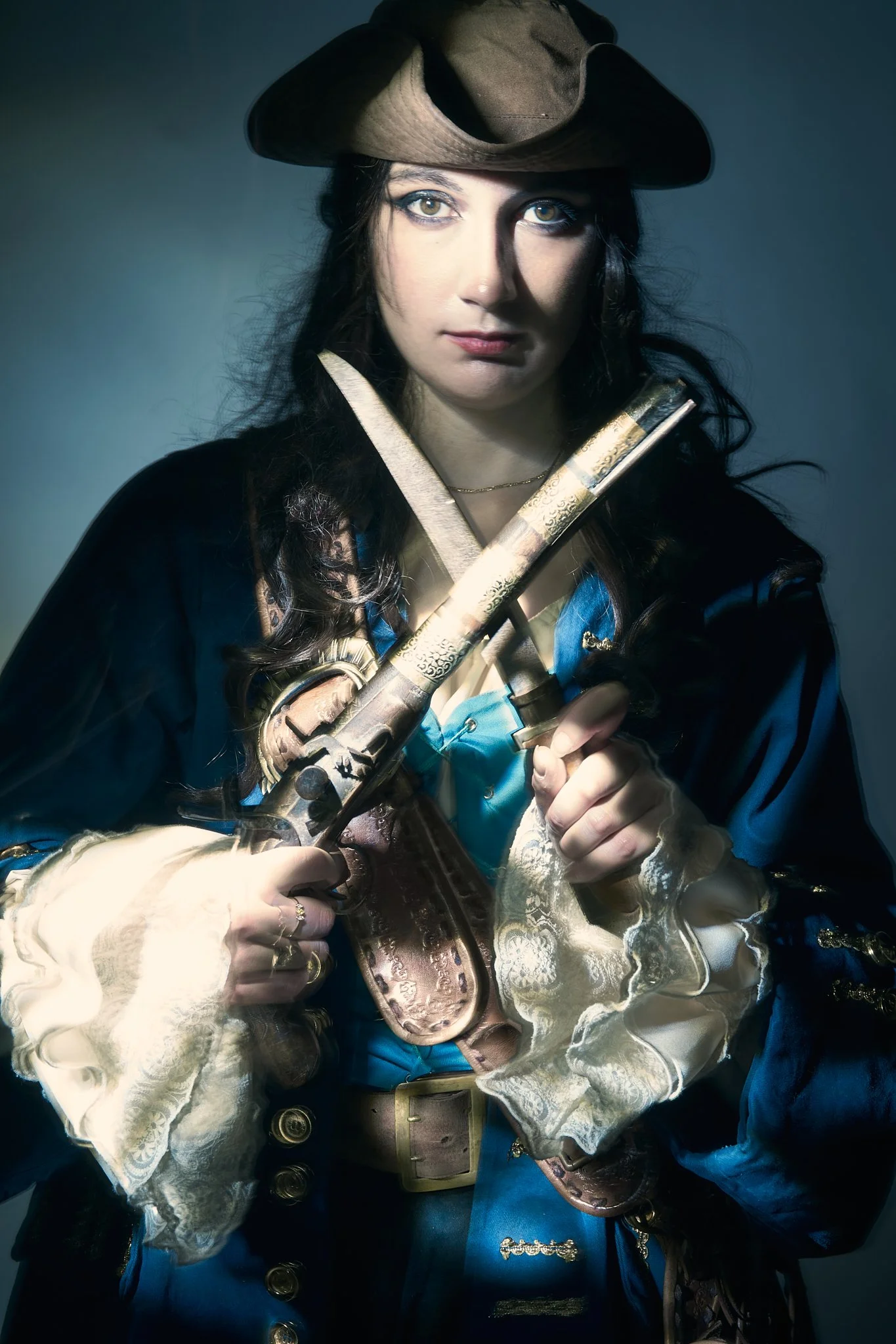 Close pirate cosplay portrait — woman in a tricorn hat and blue pirate coat with crossed sword and ornate flintlock pistol, photographed with painterly light-painting technique. Editorial cosplay photography by Simon Rogghe.