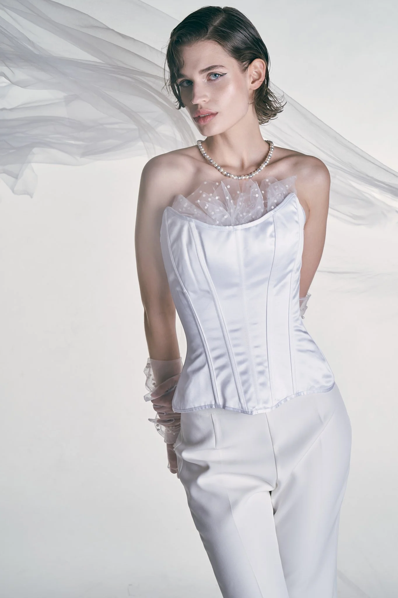 Three-quarter studio fashion photo of a model wearing a strapless white satin corset, light trousers, and sheer evening gloves under a flowing veil.⁠