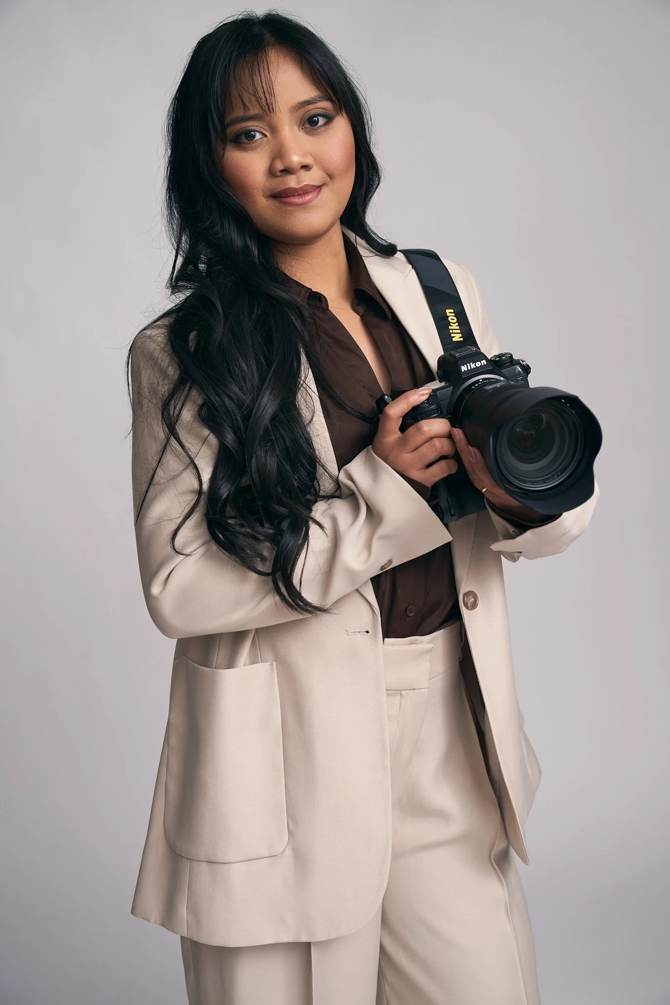 Professioneel personal branding portret van een vrouwelijke fotograaf in beige blazer, met Nikon camera in de hand. Studiofoto met zachte belichting door een portretfotograaf uit Gent, Oost-Vlaanderen.