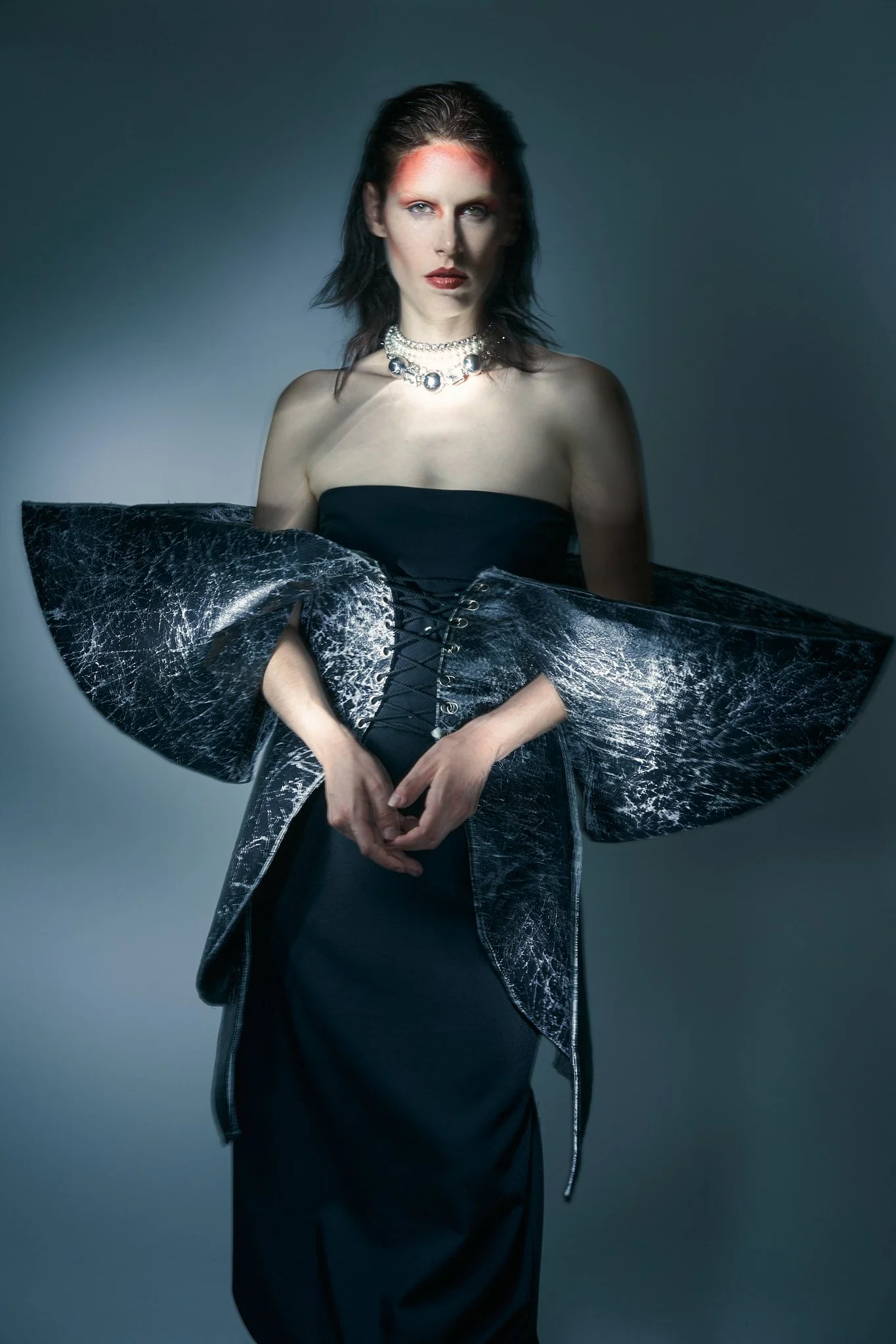 Mid-length editorial photograph of a model in a dramatic structural gown with exaggerated shoulders and modern gothic styling.⁠