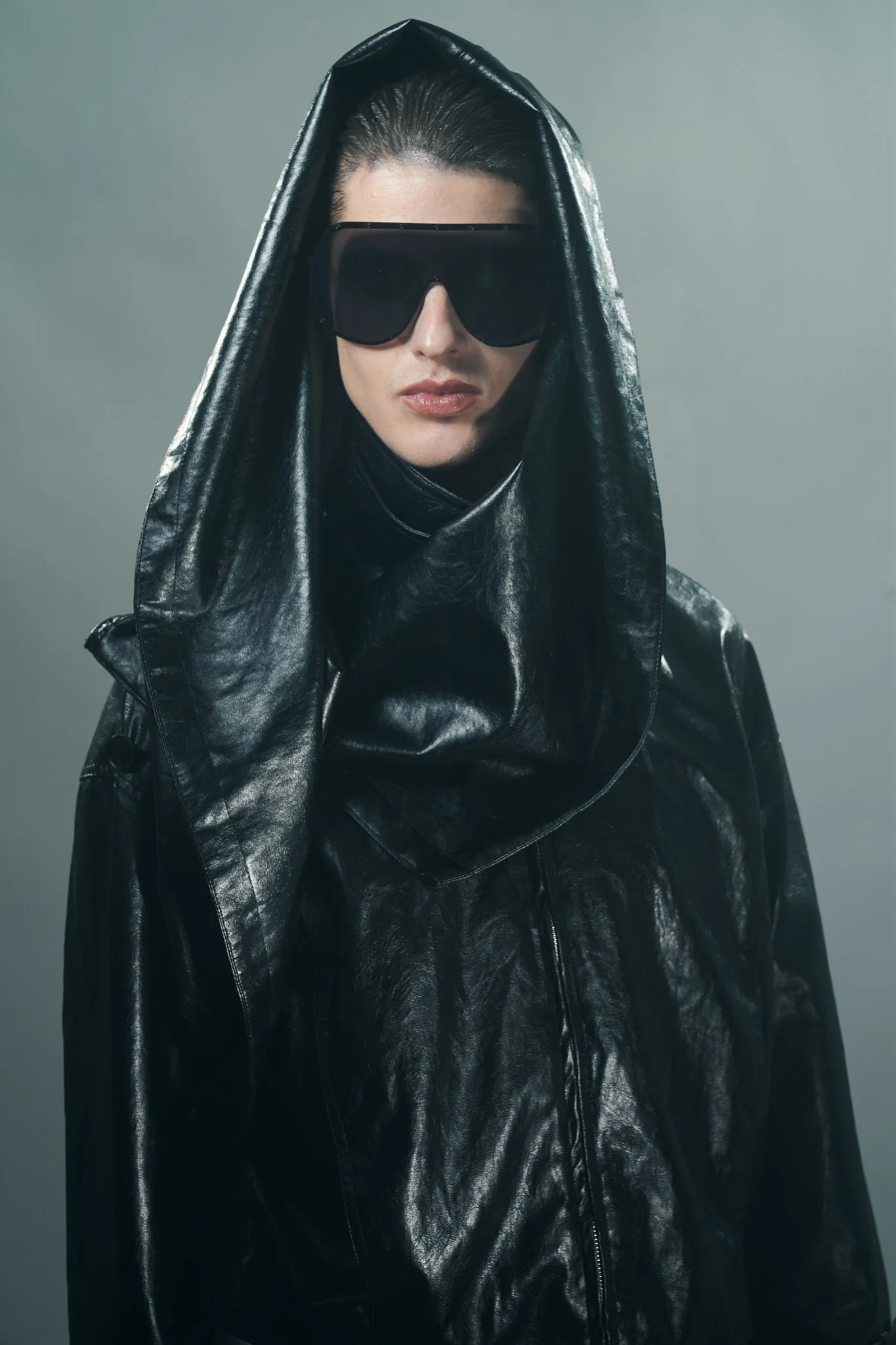 Close-up portrait of a model wearing black shield sunglasses under a dramatic oversized dark leather hood.⁠