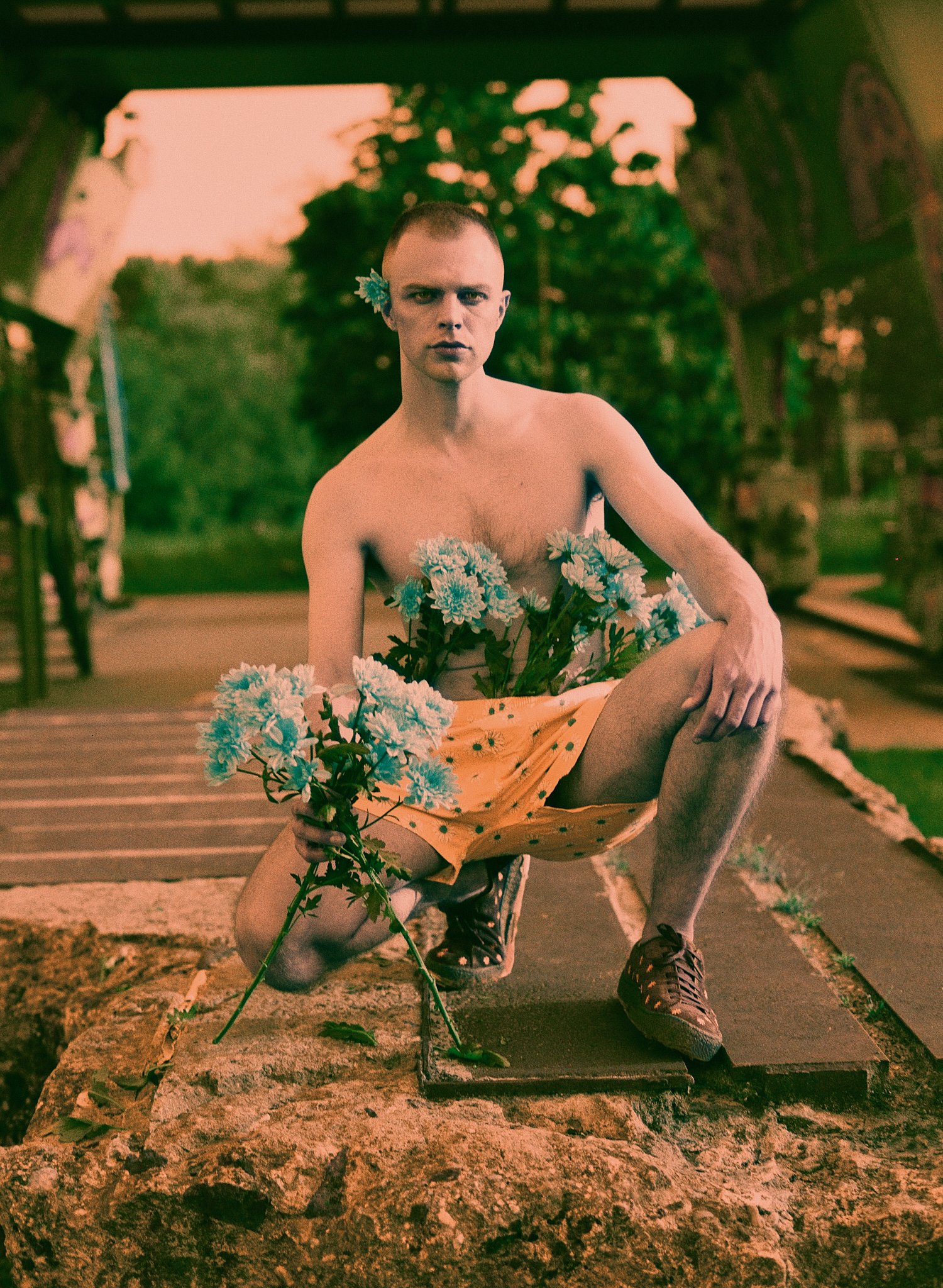 Fashion model posing outdoors with blue flowers arranged across the body in a conceptual editorial portrait featuring warm tones and artistic styling. Analog photography medium format shot on Harman Switch Azure 125 with the Mamiya 645.