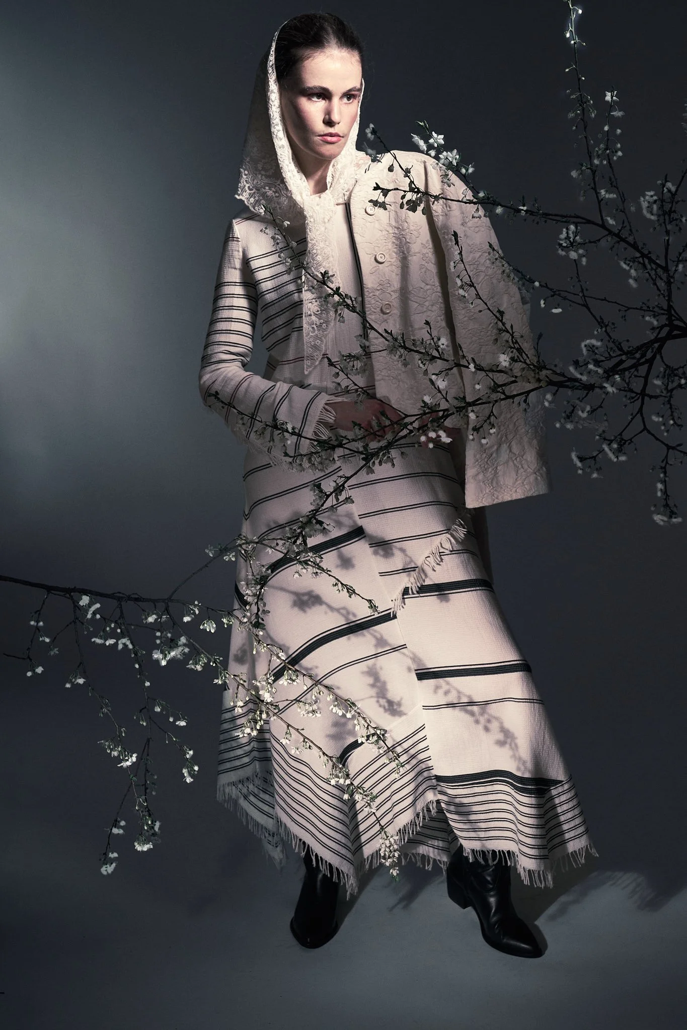 Full-body studio fashion photo of a model in a hooded cream-and-black striped knit skirt set standing beside dark floral branches.⁠