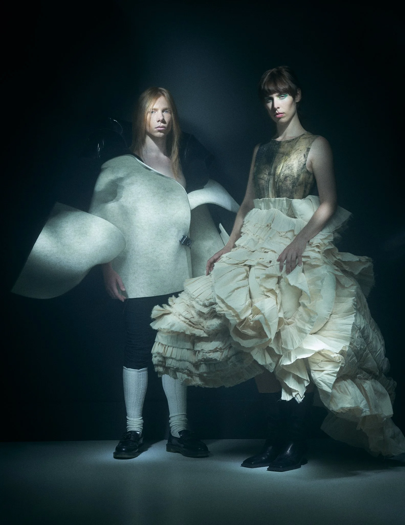Avant-garde editorial photo of two models against a dark backdrop; the woman wears an off-white tiered ruffle gown with a structured bodice, and the man wears a sculptural white felt cape silhouette paired with white socks and dark loafers.