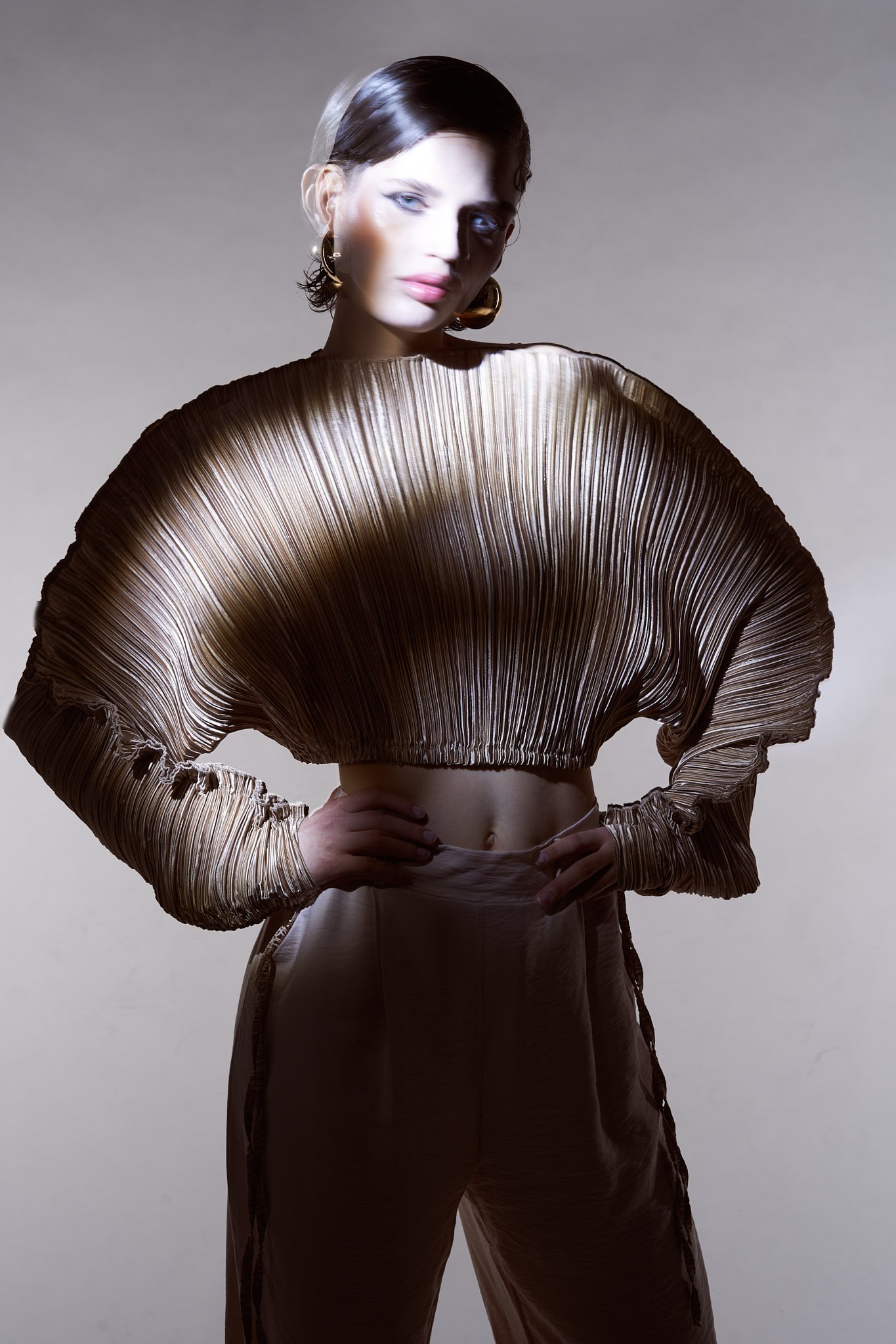 Full-length studio shot of a model wearing a wide, pleated fan-shaped crop top with matching high-waisted trousers.⁠