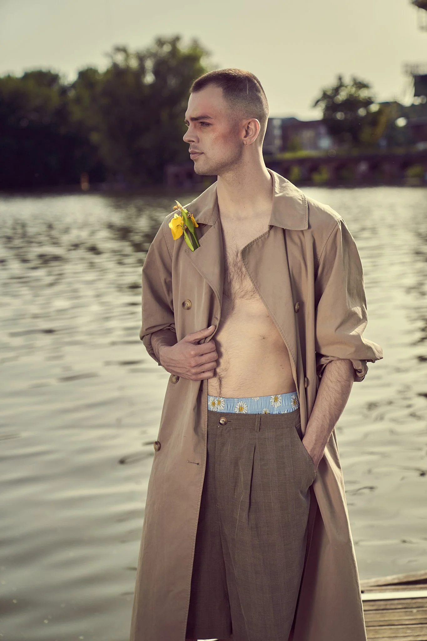 A young man in an open trench coat with a yellow flower at his lapel stands before a water backdrop, from the Animodels series by Simon Rogghe.