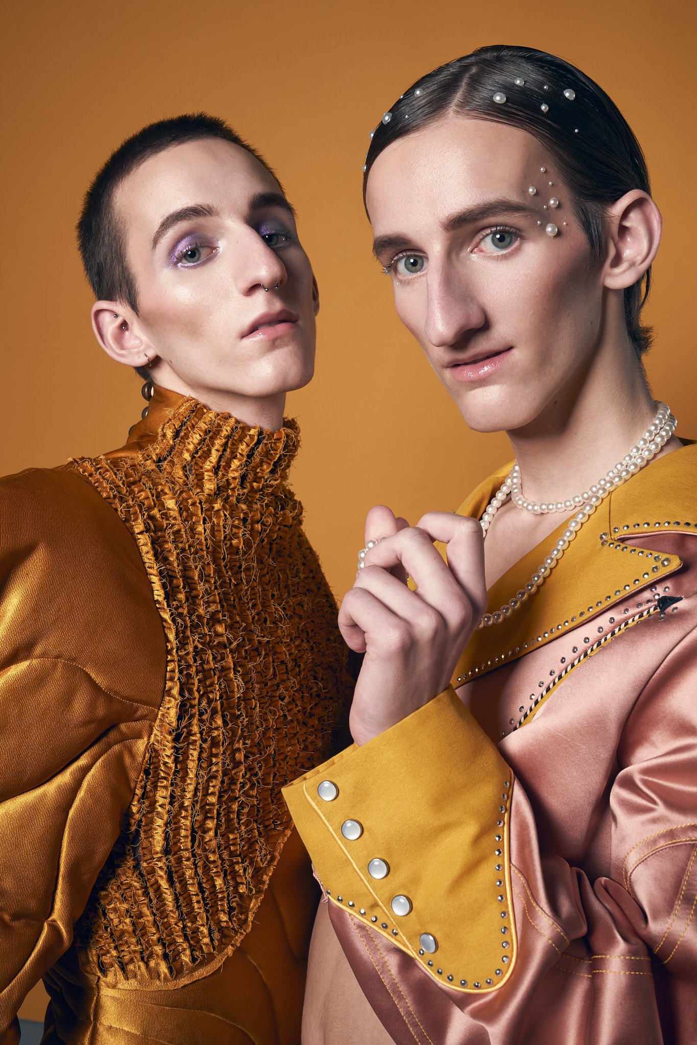 Fashion editorial portrait of twin male models in ornate styling: one wearing a textured gold high-neck top and the other in a pink jacket with pearl hair embellishments.⁠