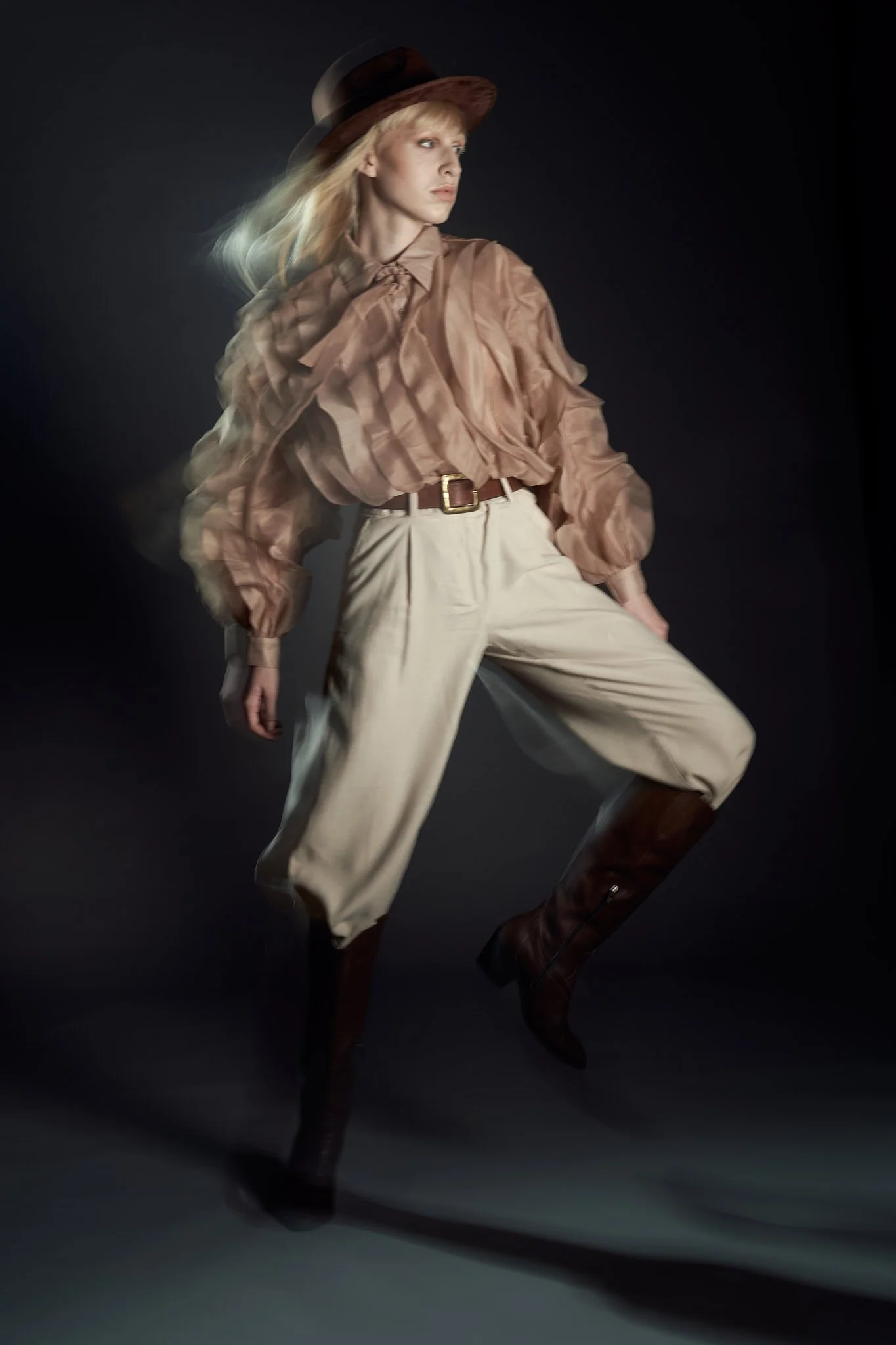 Full-body studio editorial shot of a model wearing a brown fedora, voluminous sheer beige blouse, cream riding pants, and leather boots.⁠
