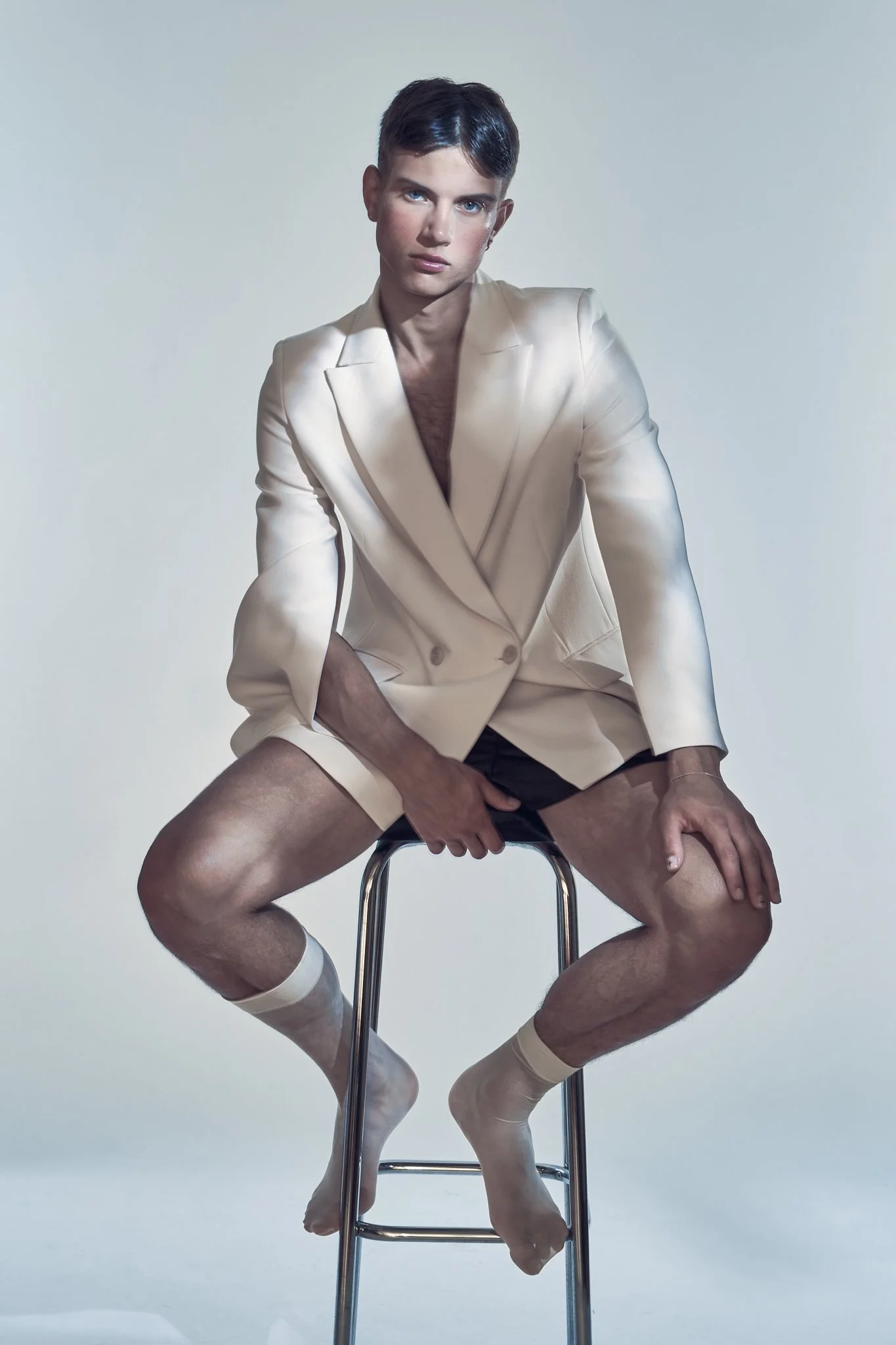 Studio portrait of a male model sitting on a stool wearing an off-white double-breasted suit jacket, dark shorts, and sheer socks.