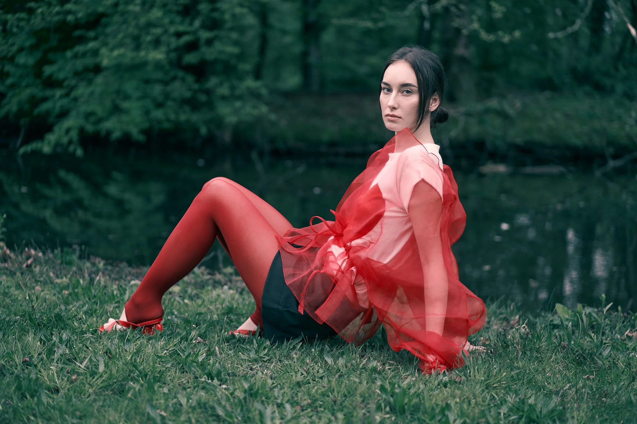 Editorial fashion portrait of a female model wearing a sheer red organza overlay and soft pink top, posed beside a lake in a natural outdoor setting, photographed for a high-fashion editorial shoot in Ghent, Belgium.