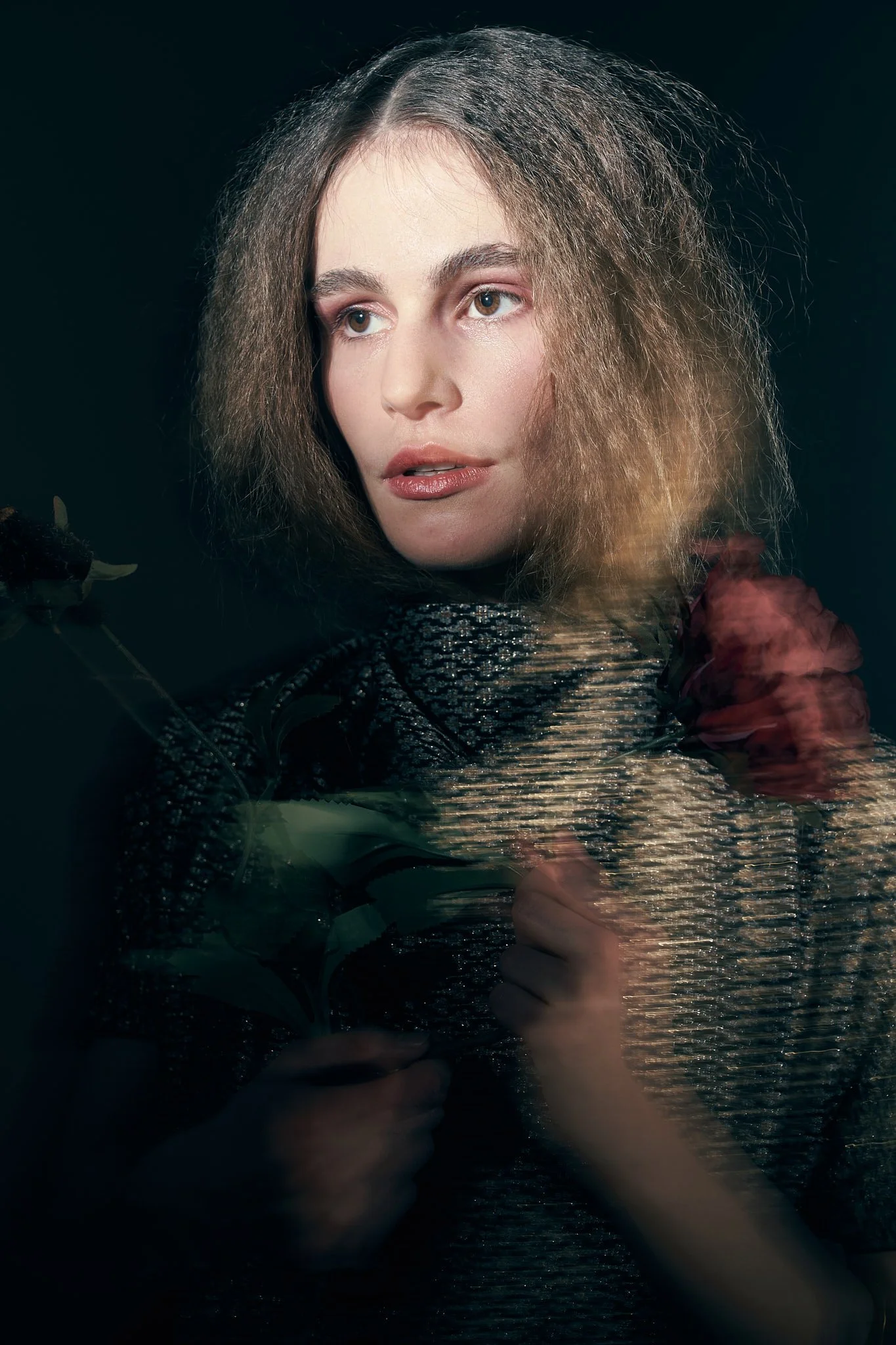 Atmospheric fashion portrait of a model with crimped hair and a red rose accent, wearing a high-neck patterned top under moody lighting.⁠