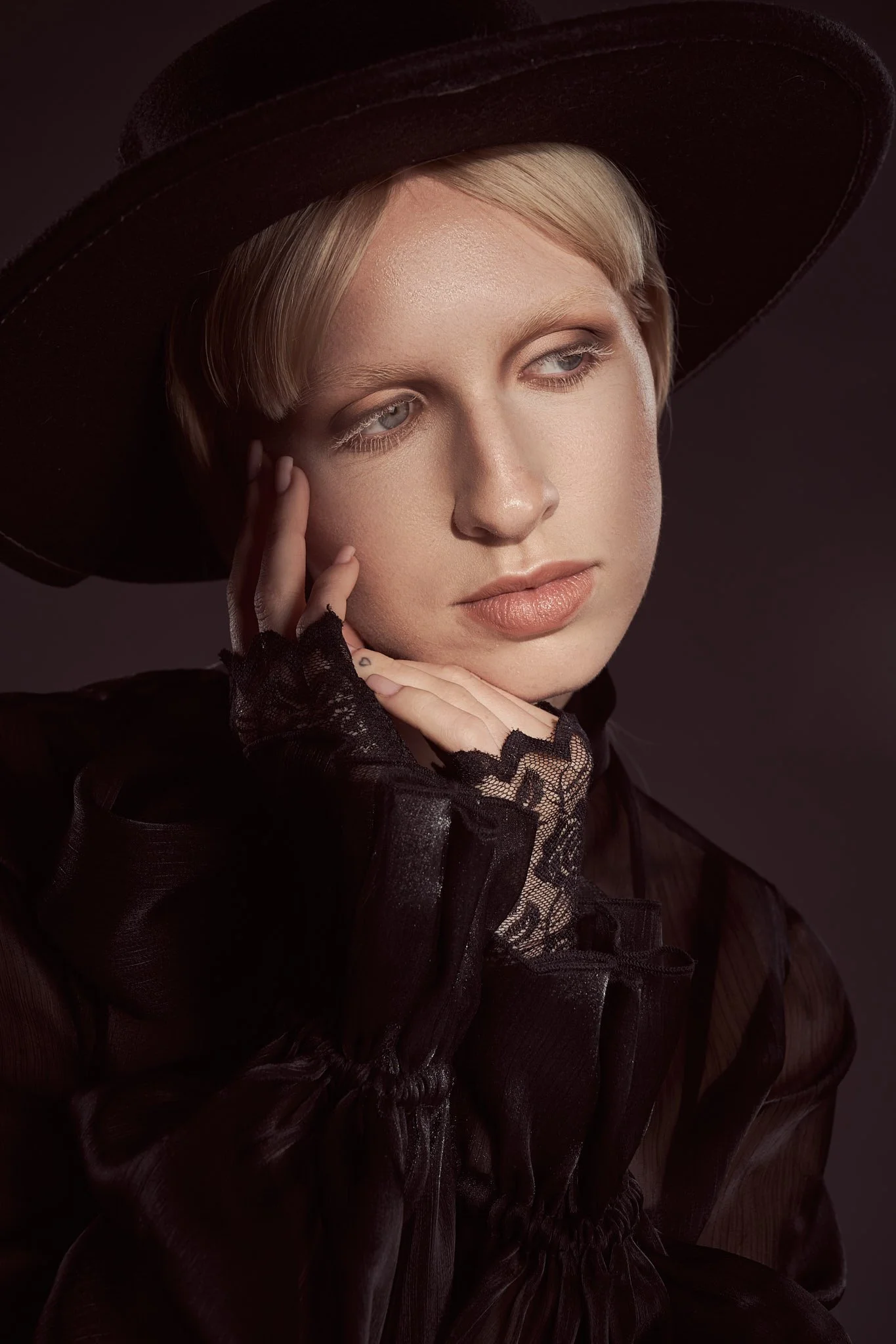 Tight close-up portrait of a blonde model with bleached eyebrows wearing a wide-brim dark hat and lace-trimmed gloves.