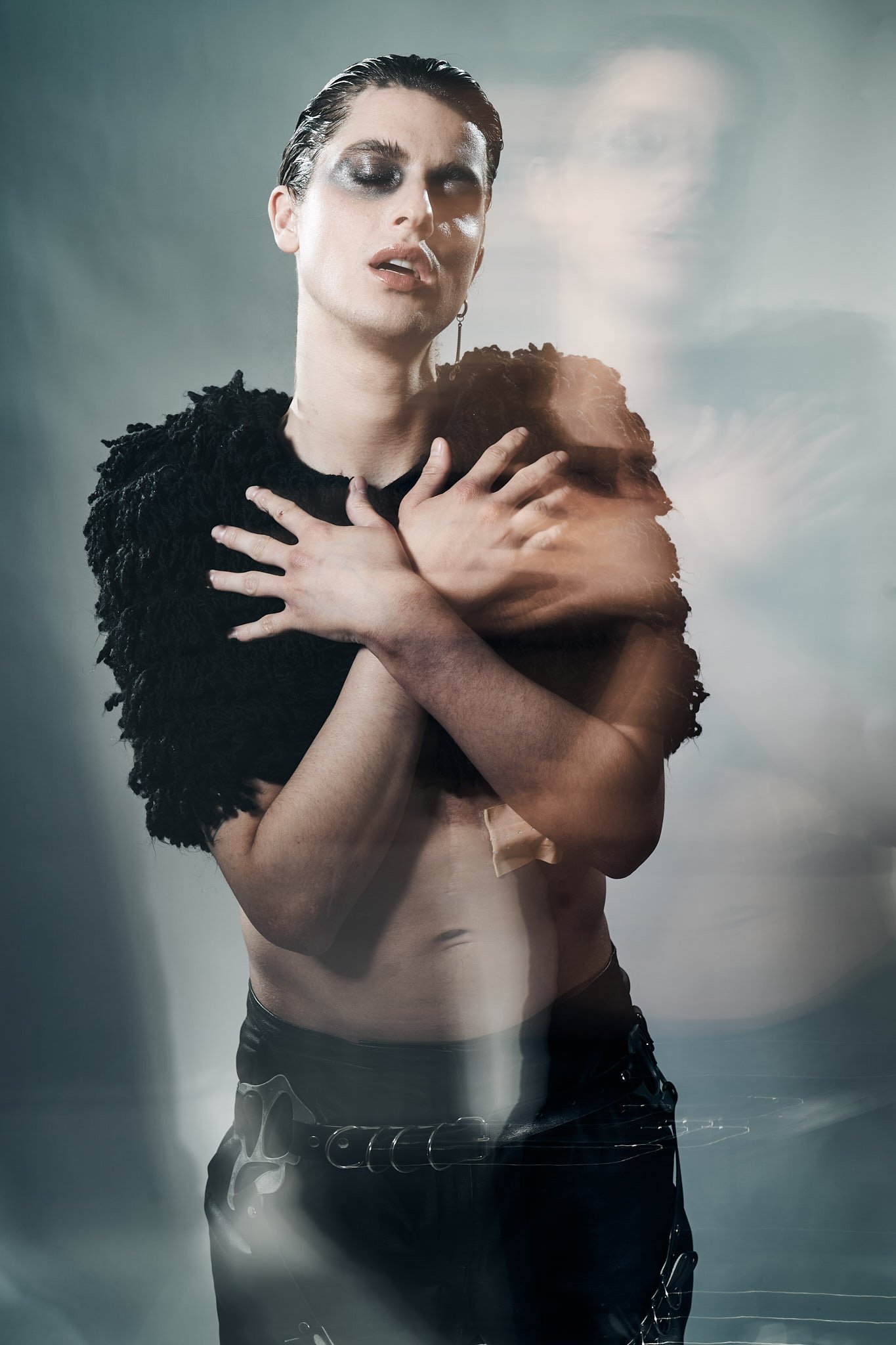Fashion portrait of a male model with slicked-back hair and smoky eye makeup, wearing a textured black top while crossing hands over chest, with soft motion blur.