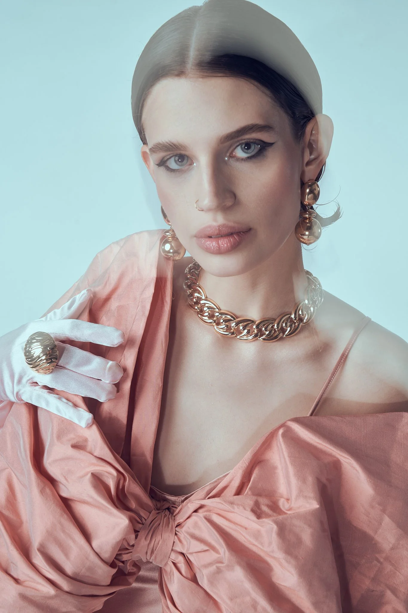 Portrait of a model wearing a thick light headband, gold chain choker, and a soft pink top against a pale blue background.⁠