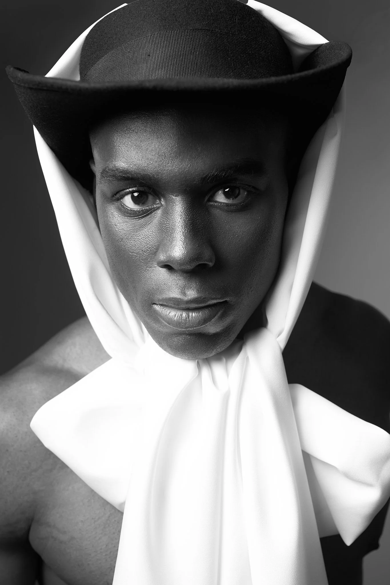 Close-up black and white portrait of a Black male model wearing a unique structural black hat and a flowing white silk neck sash.⁠
