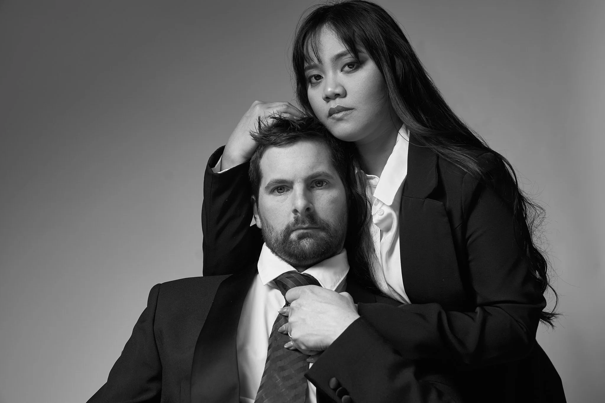 Intiem zwart-wit portret van een koppel tijdens een loveshoot in Gent, Oost-Vlaanderen. De vrouw zit dicht bij haar partner, houdt zijn das vast en rust haar hand zacht in zijn haar. Een elegante en artistieke koppelshoot met cinematografische sfeer.