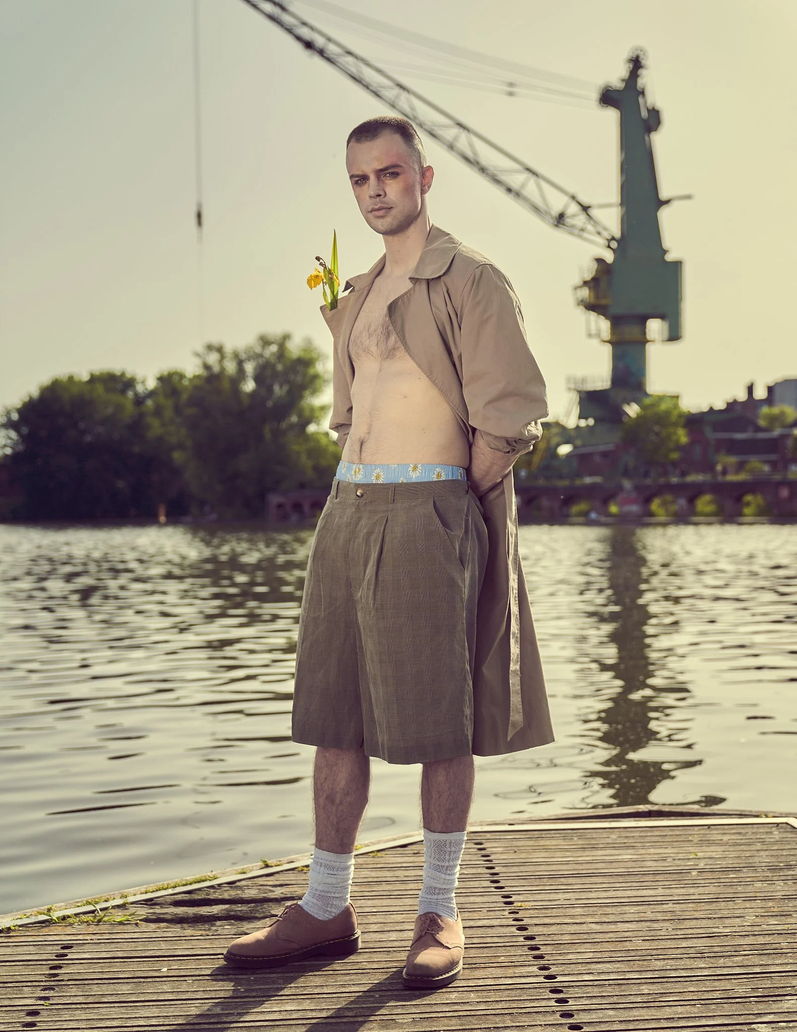 Editorial fashion portrait of a young man in an open terracotta wool coat with floral brooches, crouching among green foliage at an industrial waterfront location in Ghent, Belgium.