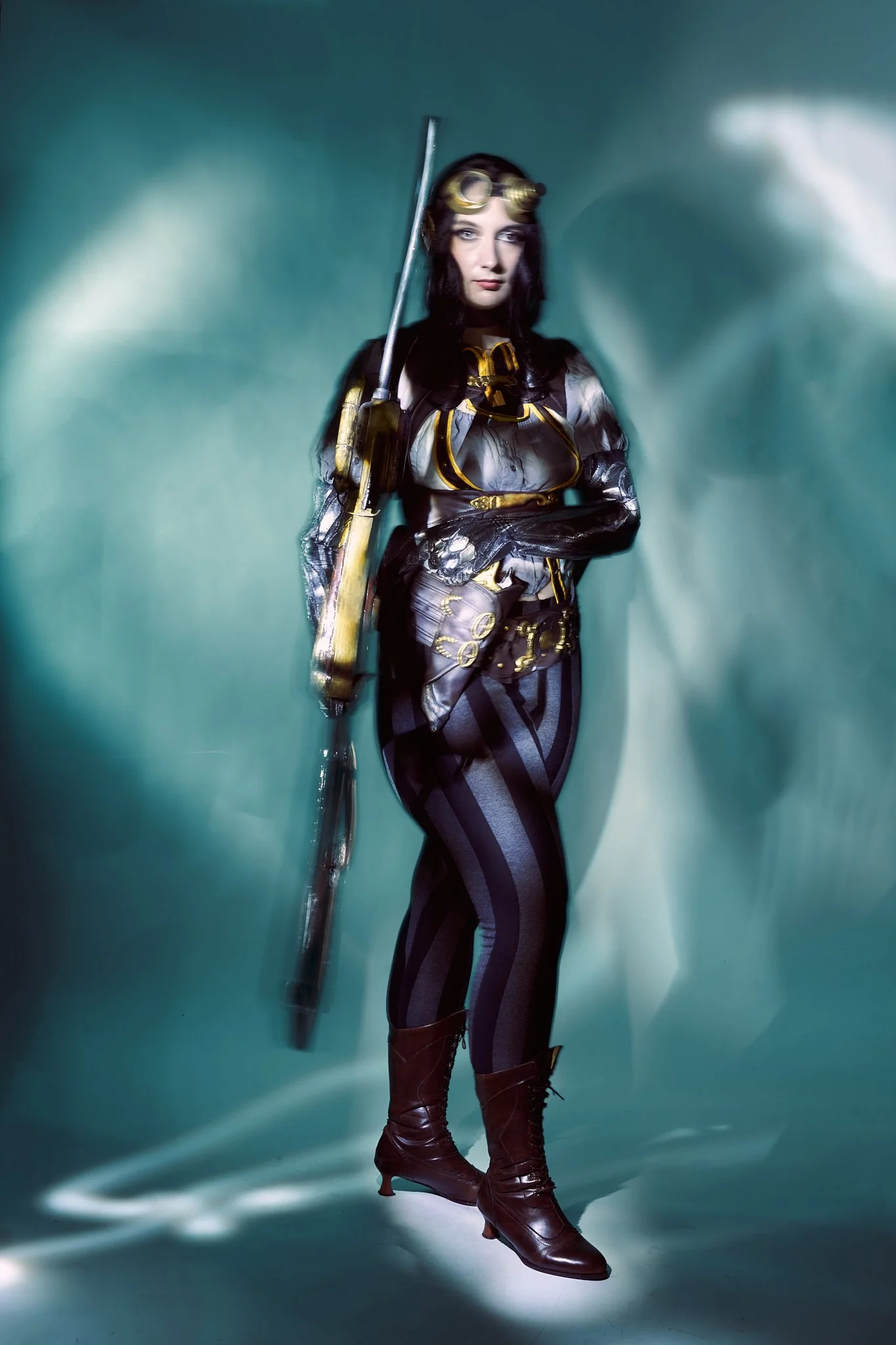 Full-body Lady Mechanika cosplay portrait — woman in brass goggles, black-and-gold harnessed corset with leather belt and silver mechanical arm, shot with light-painting technique on teal. Editorial photography by Simon Rogghe.