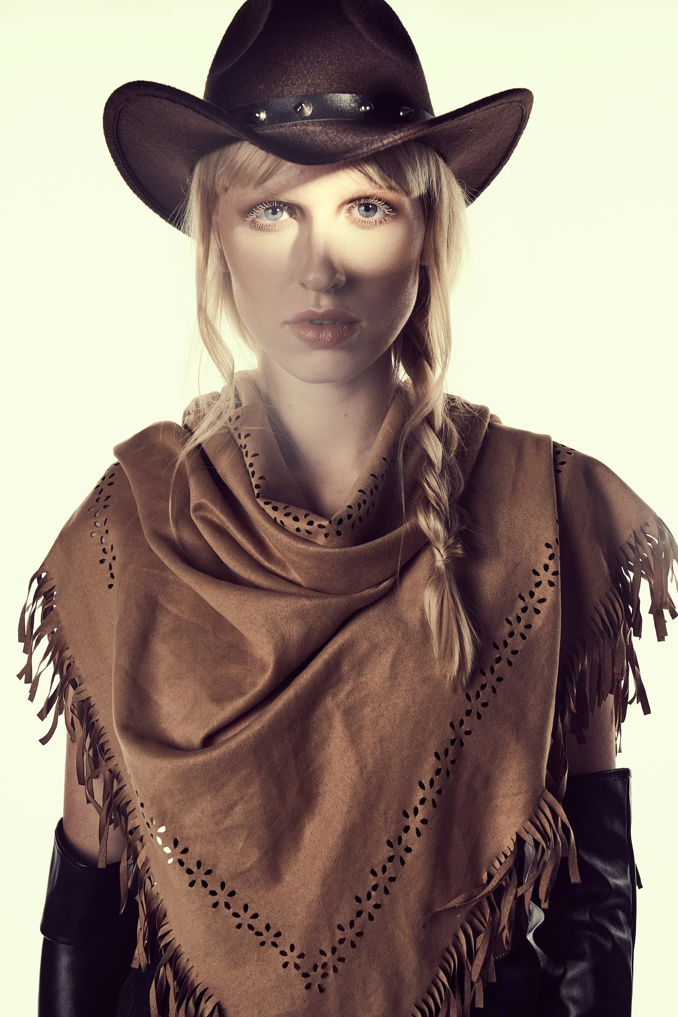 Close-up portrait of a blonde model with a side braid wearing a studded leather cowboy hat and a laser-cut brown fringed shawl.⁠