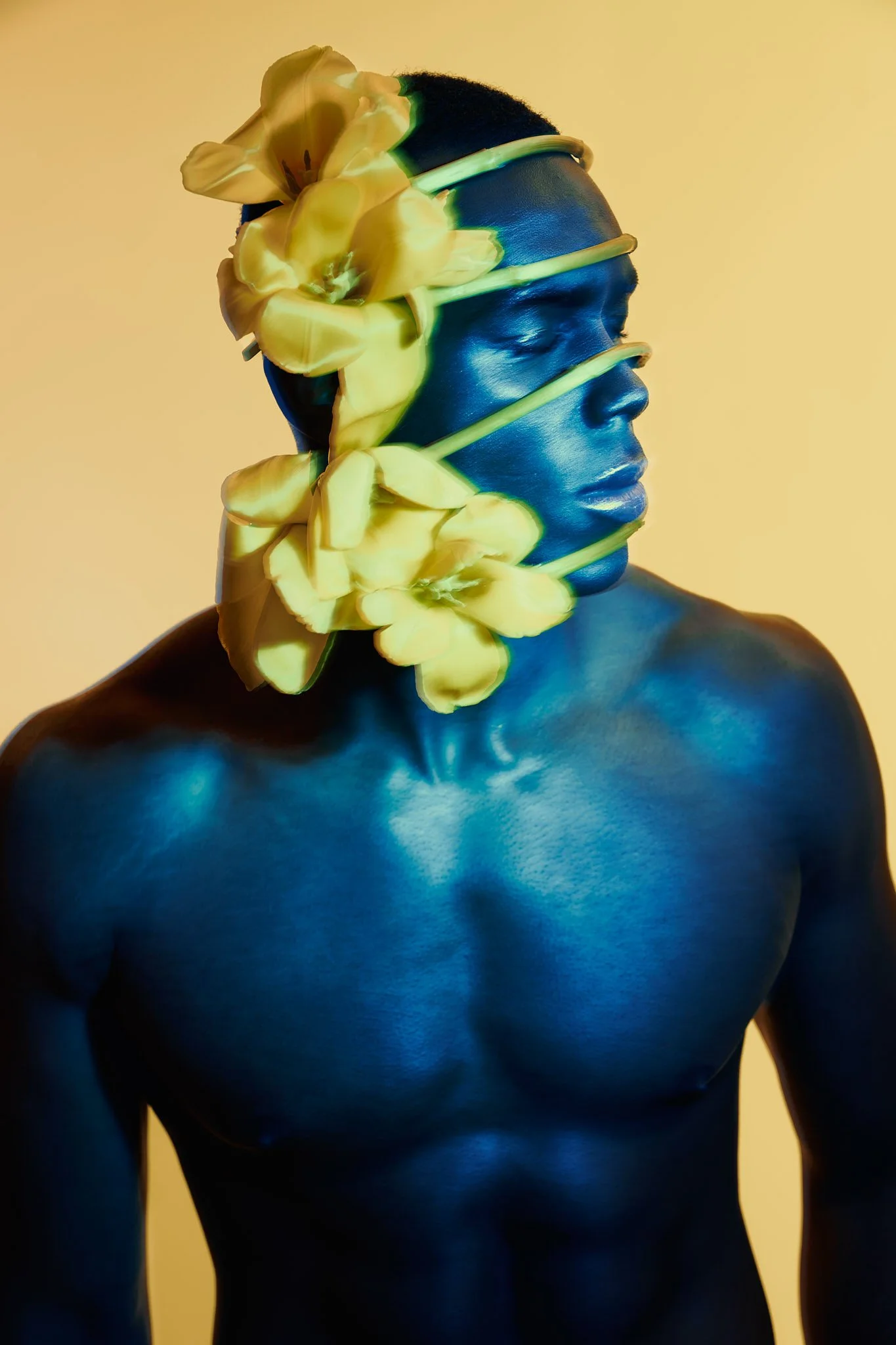 Artistic portrait of a Black male model with vibrant blue body paint covering his chest, shoulders, and face, wearing large yellow flowers as a headpiece.⁠