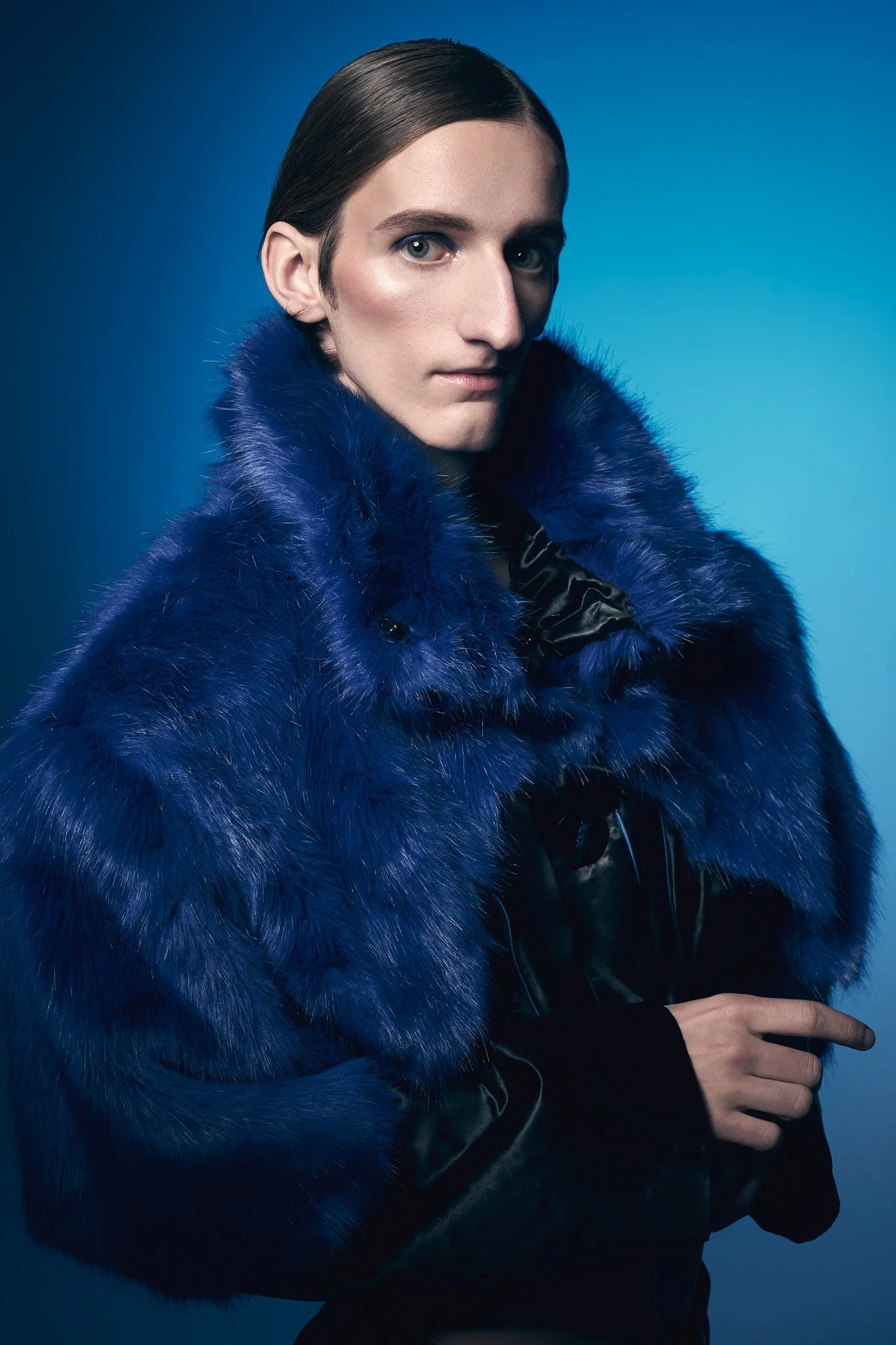 High-fashion studio portrait of a male model wearing an oversized cobalt blue faux fur jacket with a high collar against a solid blue background.⁠