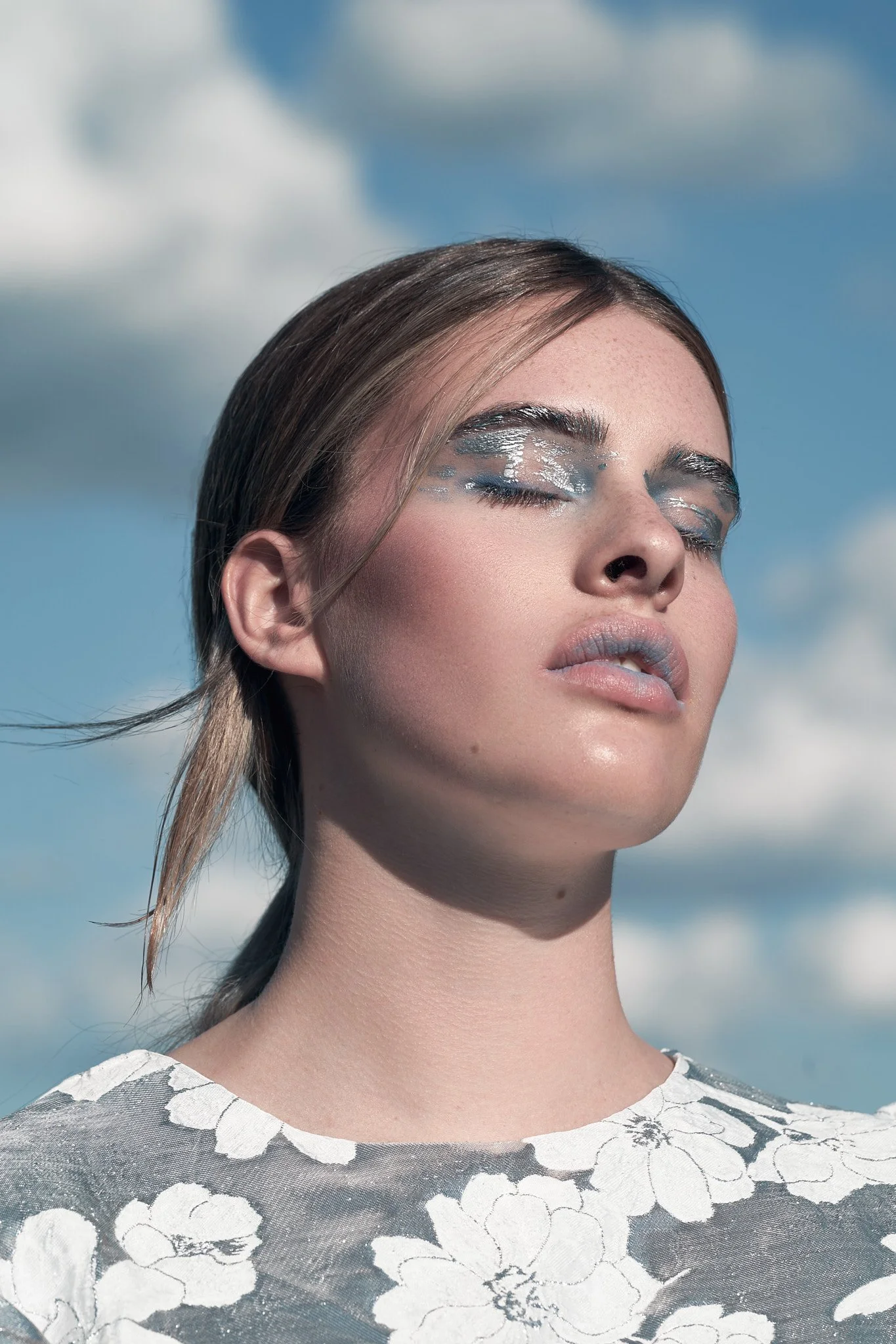 Editorial fashion portrait, model with silver makeup and eyes closed tilted toward an open sky, metallic floral dress, analog photography