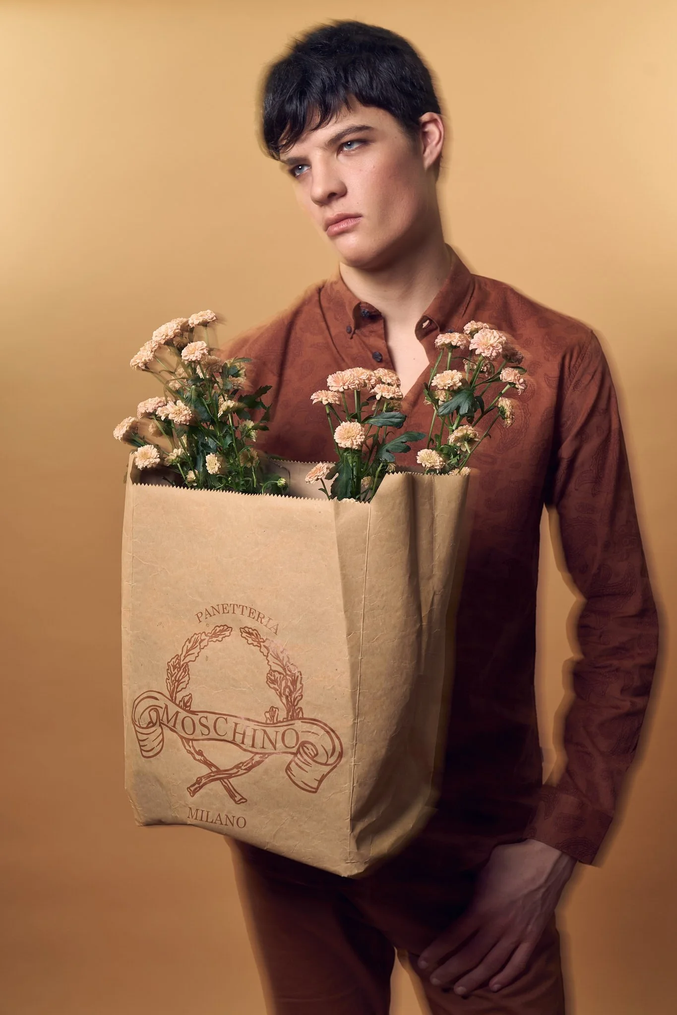 Editorial fashion portrait of an androgynous model holding a Moschino paper bag with flowers, photographed in studio by a Belgian fashion photographer.