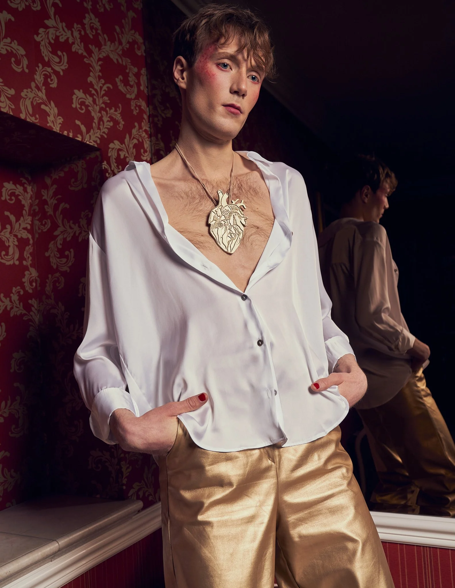 Three-quarter editorial shot of a male model in an unbuttoned white silk shirt, gold trousers, and a large heart-shaped pendant, in an ornate vintage room.⁠