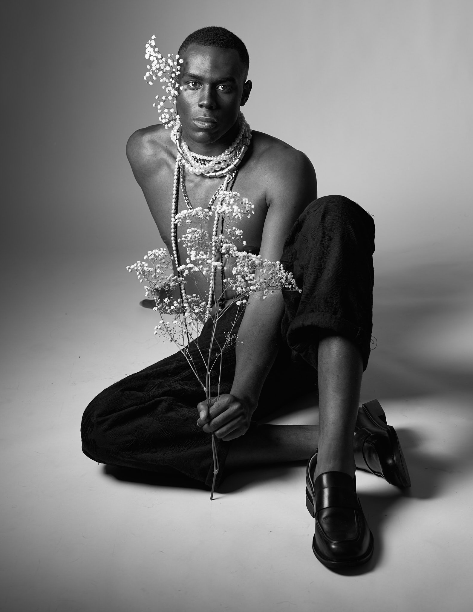 Full-body black and white photograph of a Black male model sitting cross-legged, shirtless, with layers of beaded necklaces, holding baby's breath flowers.⁠