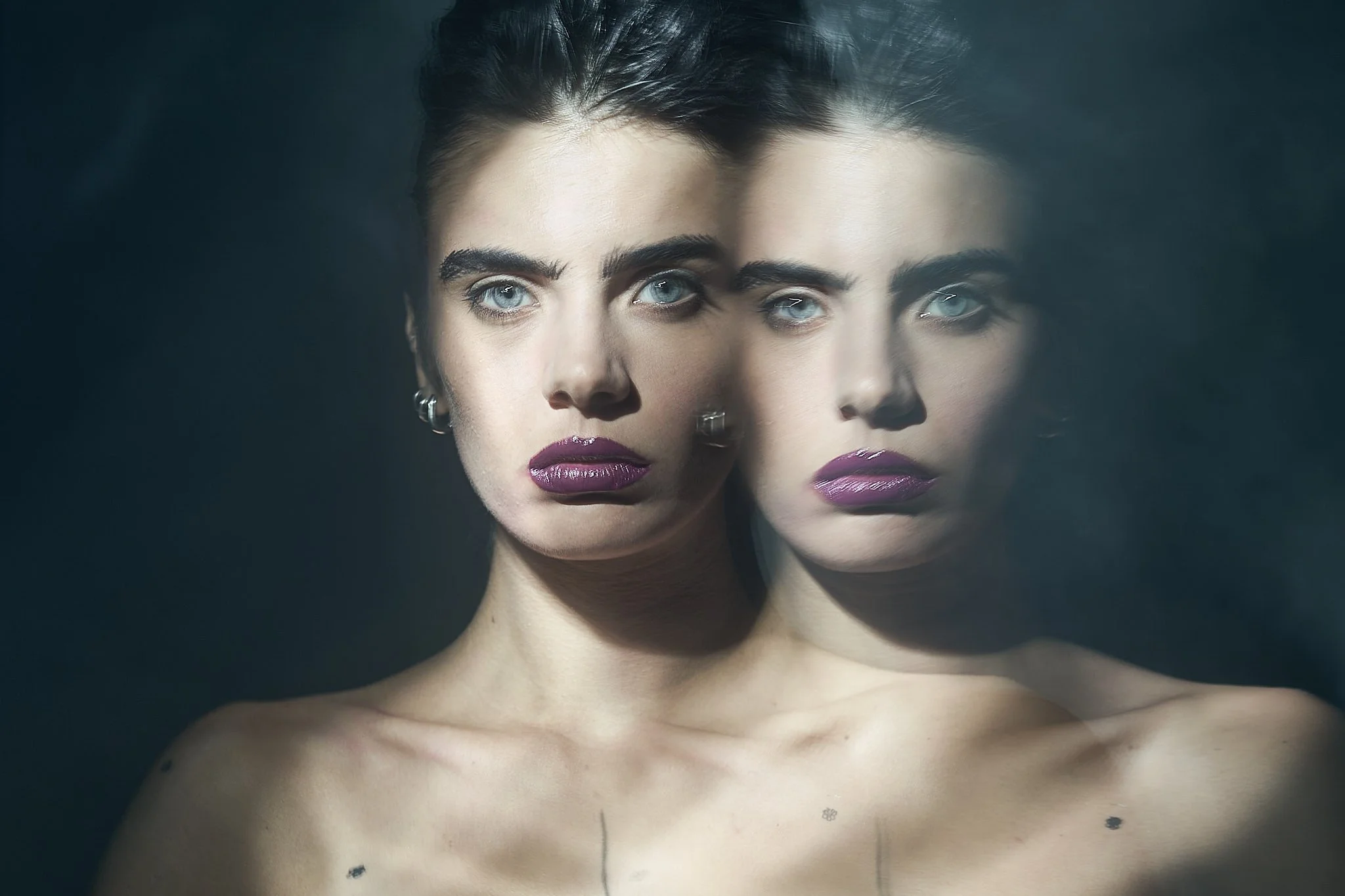 Surreal beauty editorial shot showing a dual image reflection of a model with slicked hair, defined brows, and deep berry lip color.⁠