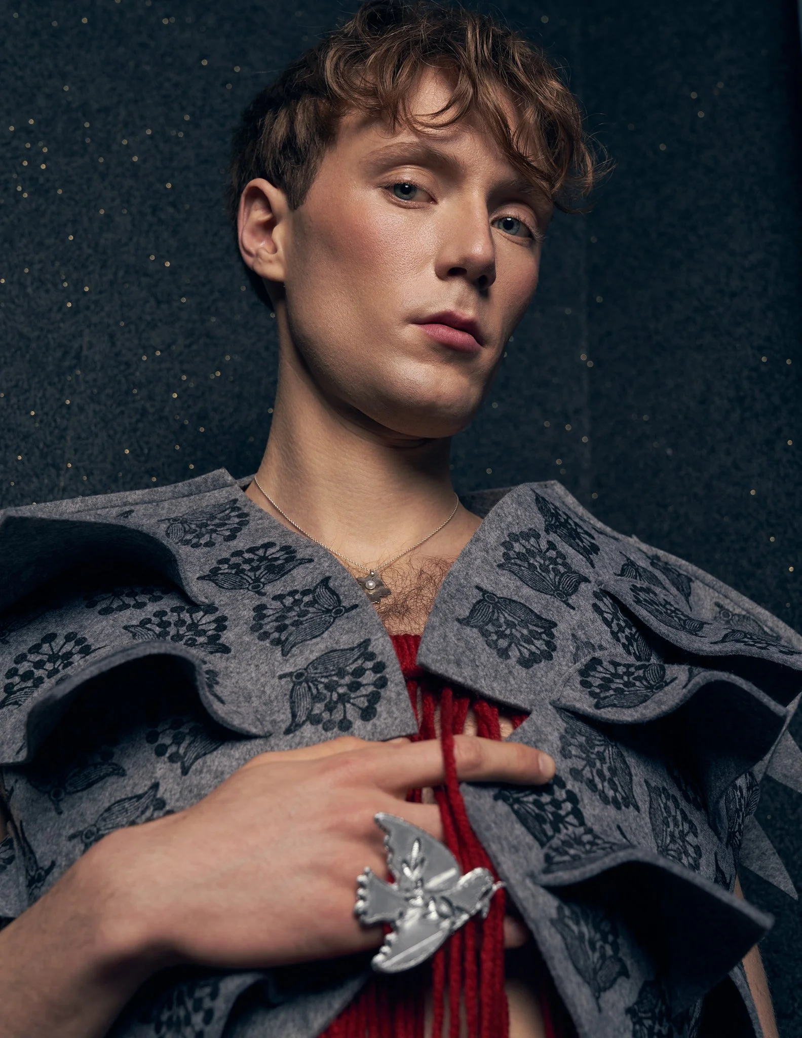 Close-up portrait of a male model wearing a grey jacket with dramatic tiered scalloped collars and a silver bird brooch.⁠